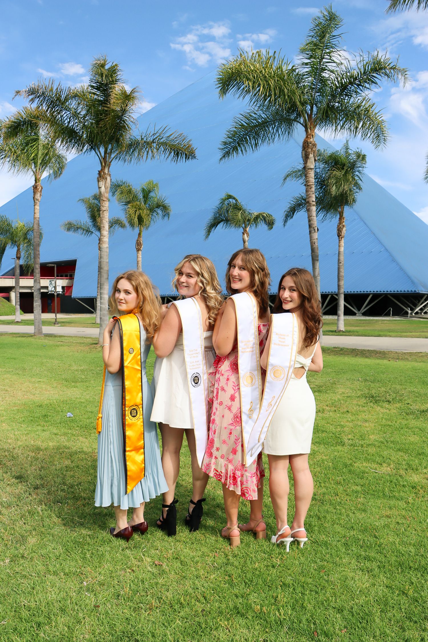 A group in summer dresses stands together on a grassy lawn with palm trees and blue pyramids in the background.