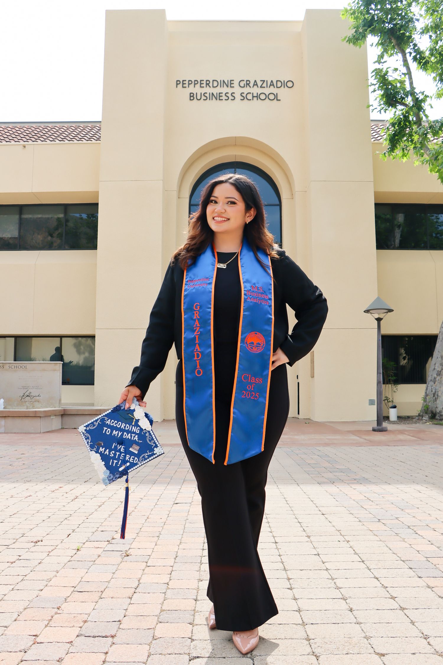 Graduate wearing a black dress and blue graduation stole with red embroidery poses outside Pepperdine Graziadio Business School.