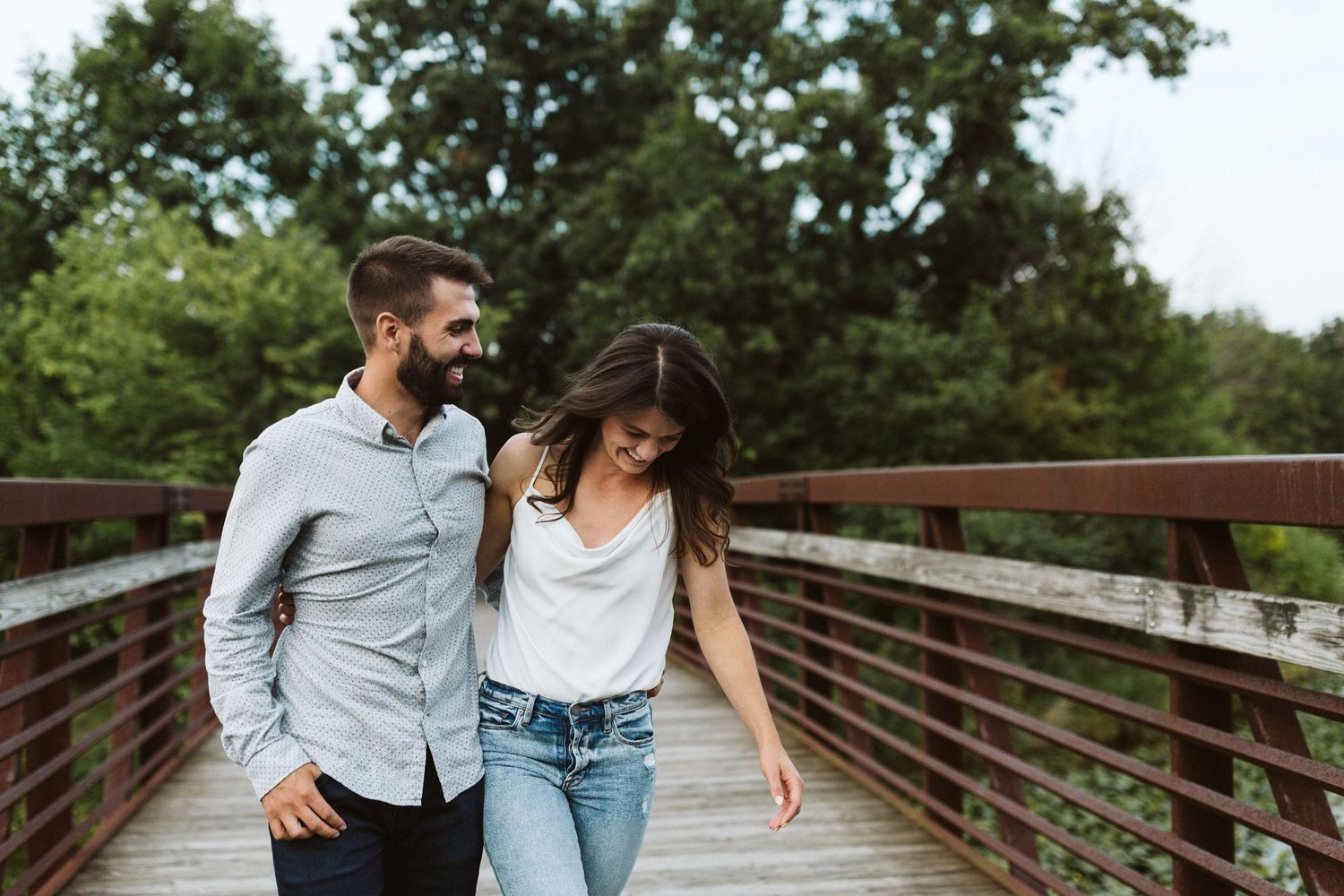 A couple in casual attire walking together on a wooden bridge in a lush green park setting.