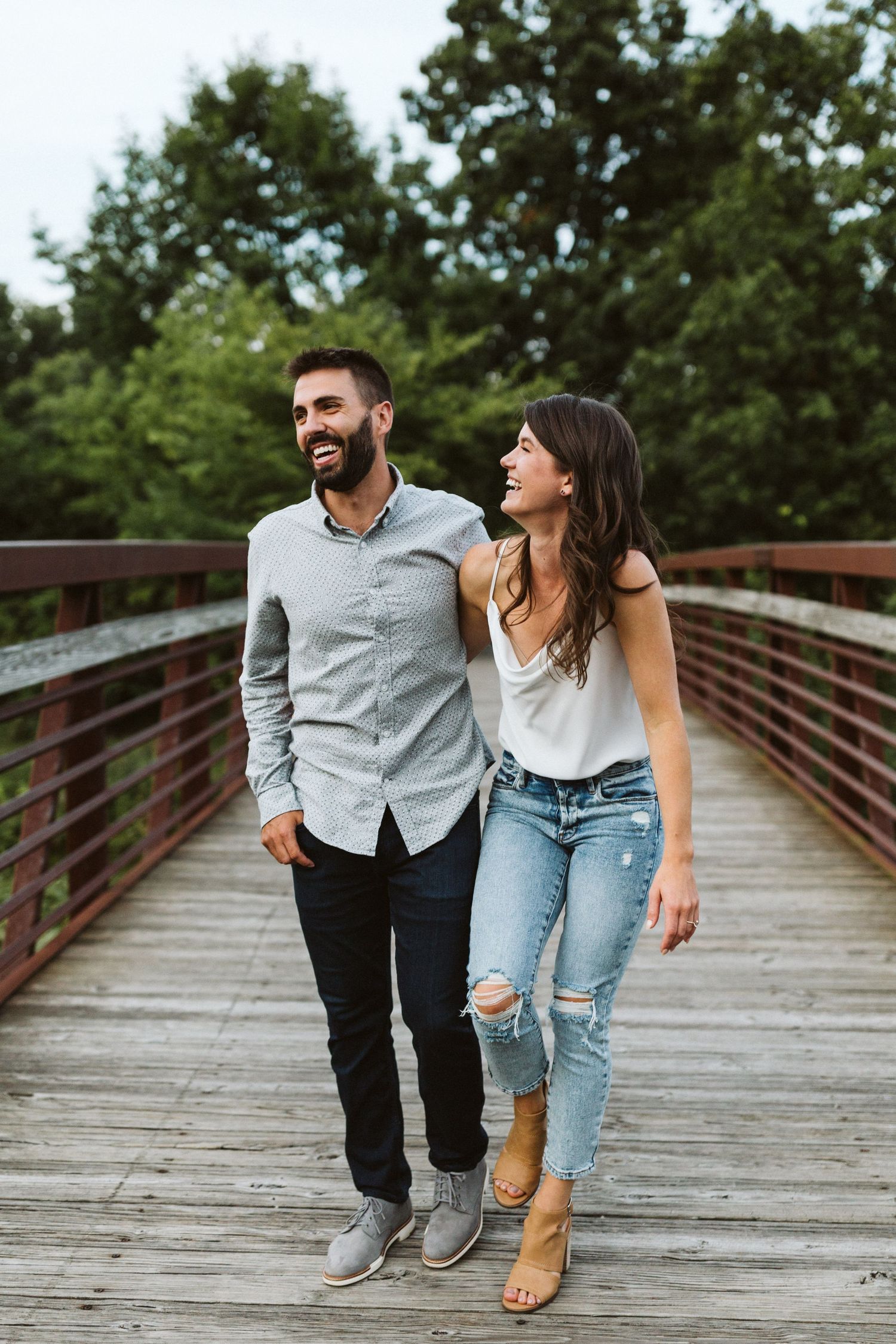 A couple laughing together while walking on a wooden footbridge in a park setting.