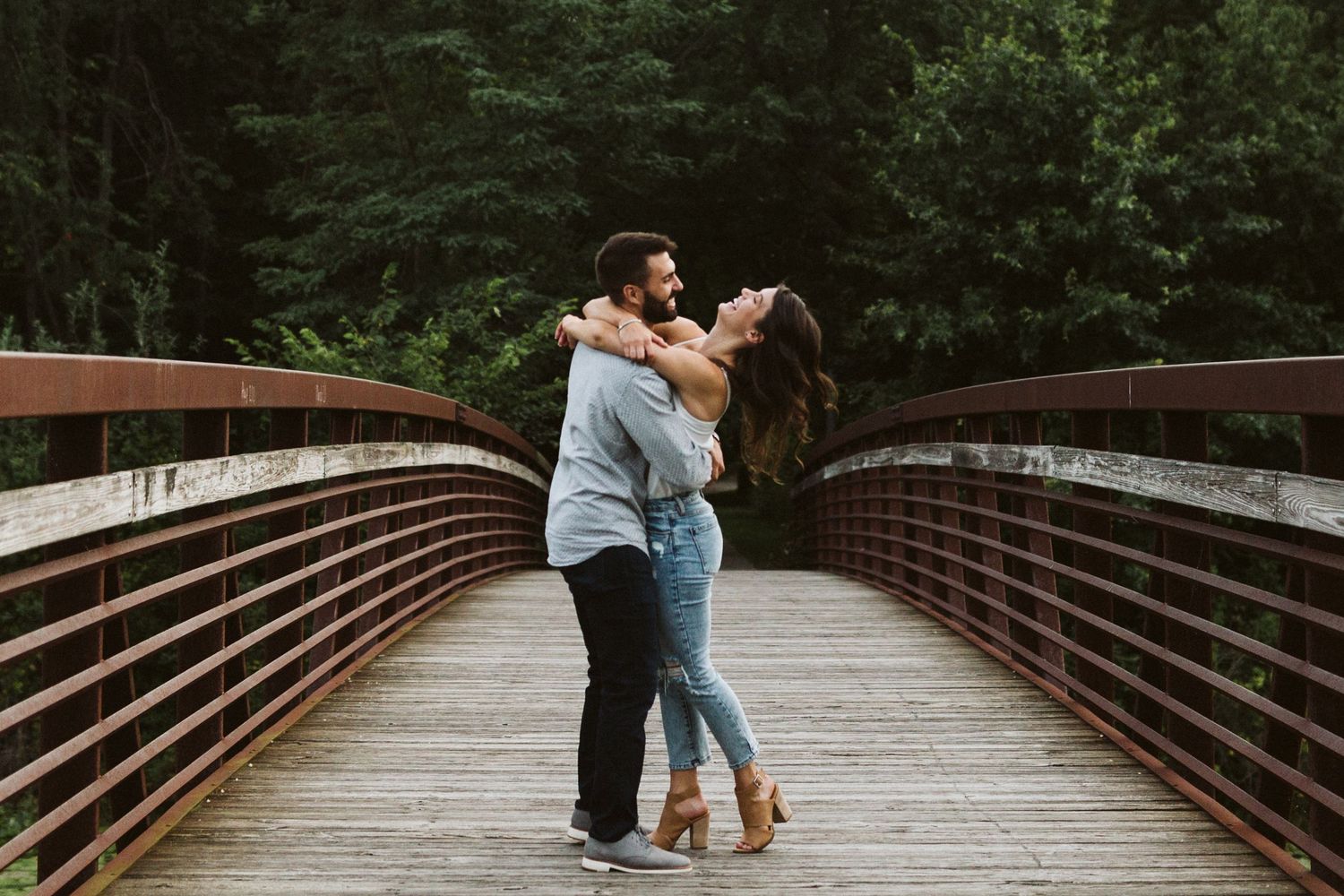 A couple embraces romantically on a wooden bridge surrounded by lush green forest scenery.