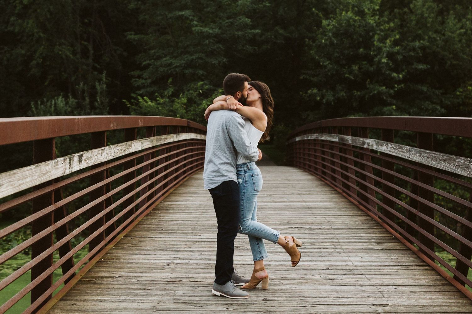 A couple embraces and shares a romantic moment on a wooden bridge surrounded by lush greenery.