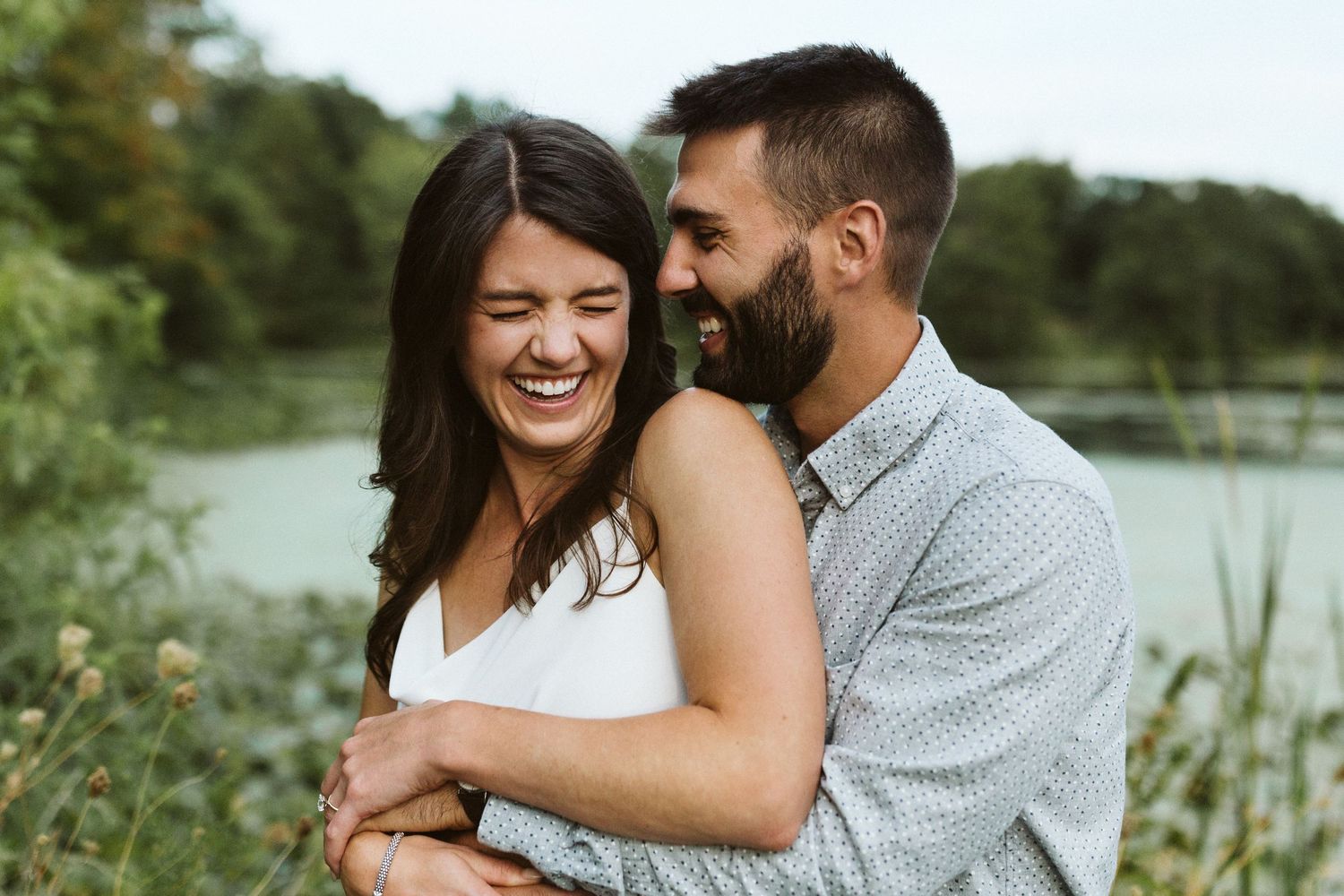 A couple shares joyful embraces and laughter during an outdoor photo session near a lake and lush greenery.