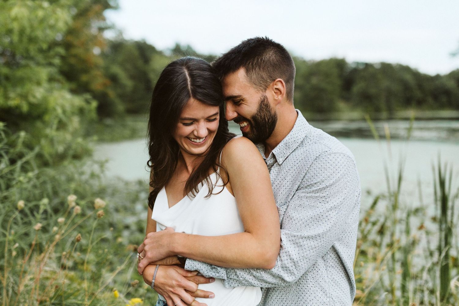 A couple shares a romantic moment near a scenic lake with lily pads and lush greenery in the background.