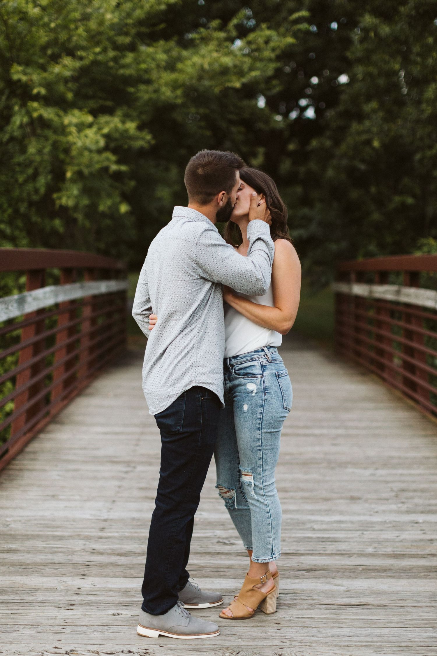 A romantic couple sharing an intimate moment while standing on a wooden bridge surrounded by green trees.