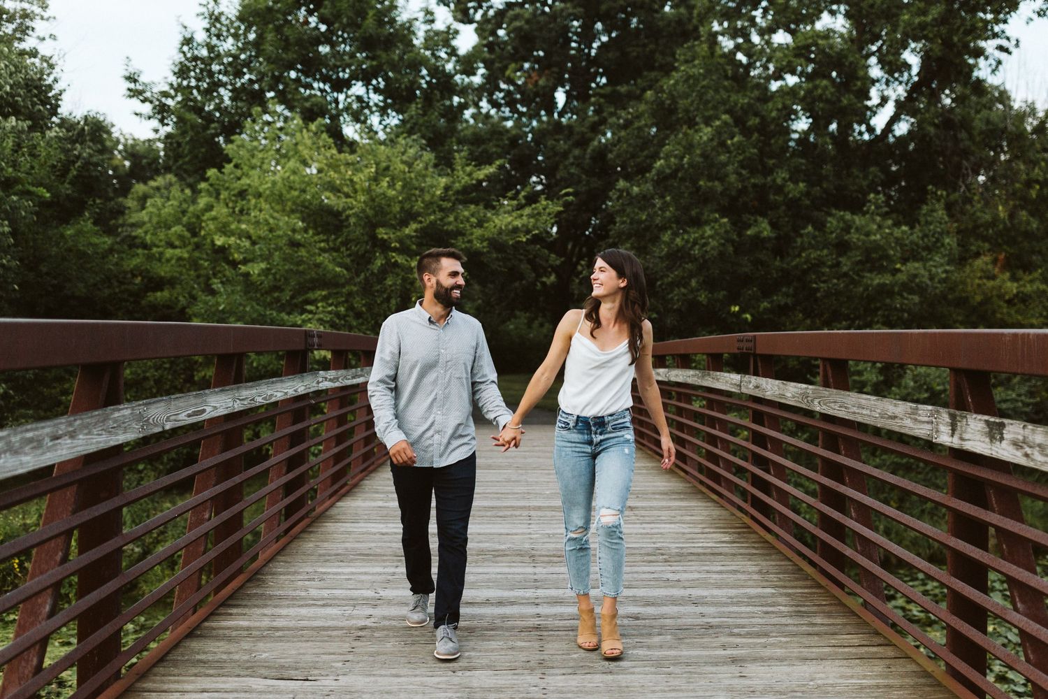 Couple walking hand in hand across a wooden bridge surrounded by lush greenery on a sunny day.