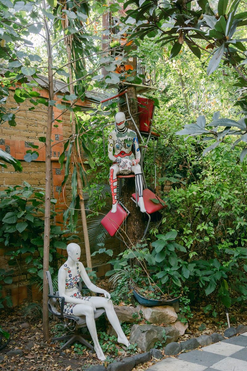 White ceramic decorative figures displayed in a lush garden setting with brick wall and greenery.