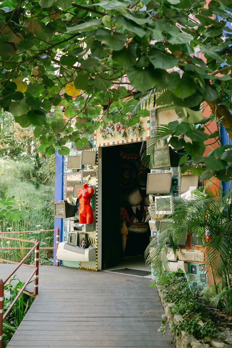 Wooden deck walkway surrounded by lush green foliage and hanging decorations in an outdoor garden setting.