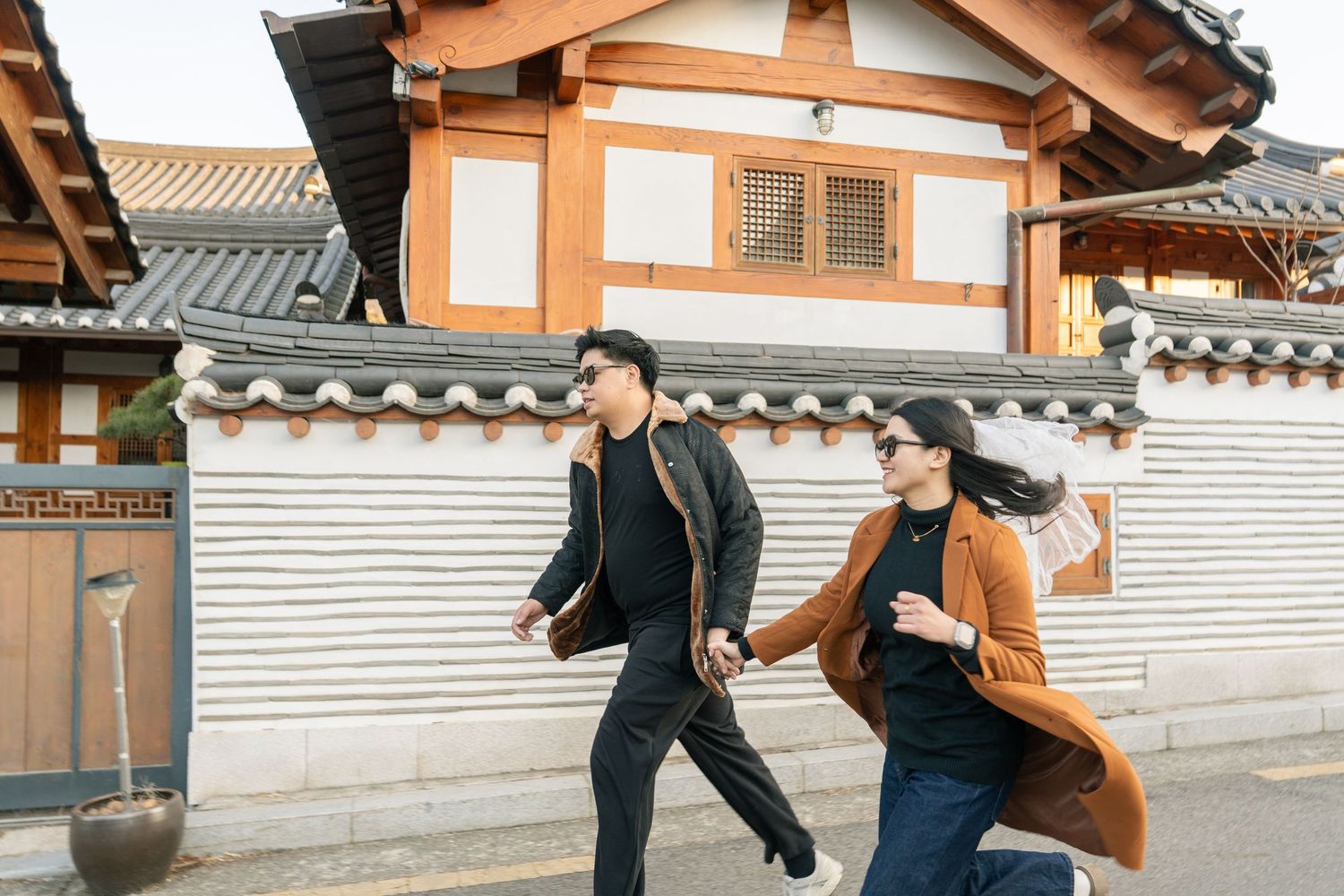 Two people in winter coats walk together past traditional Korean hanok architecture with tiled roofs.