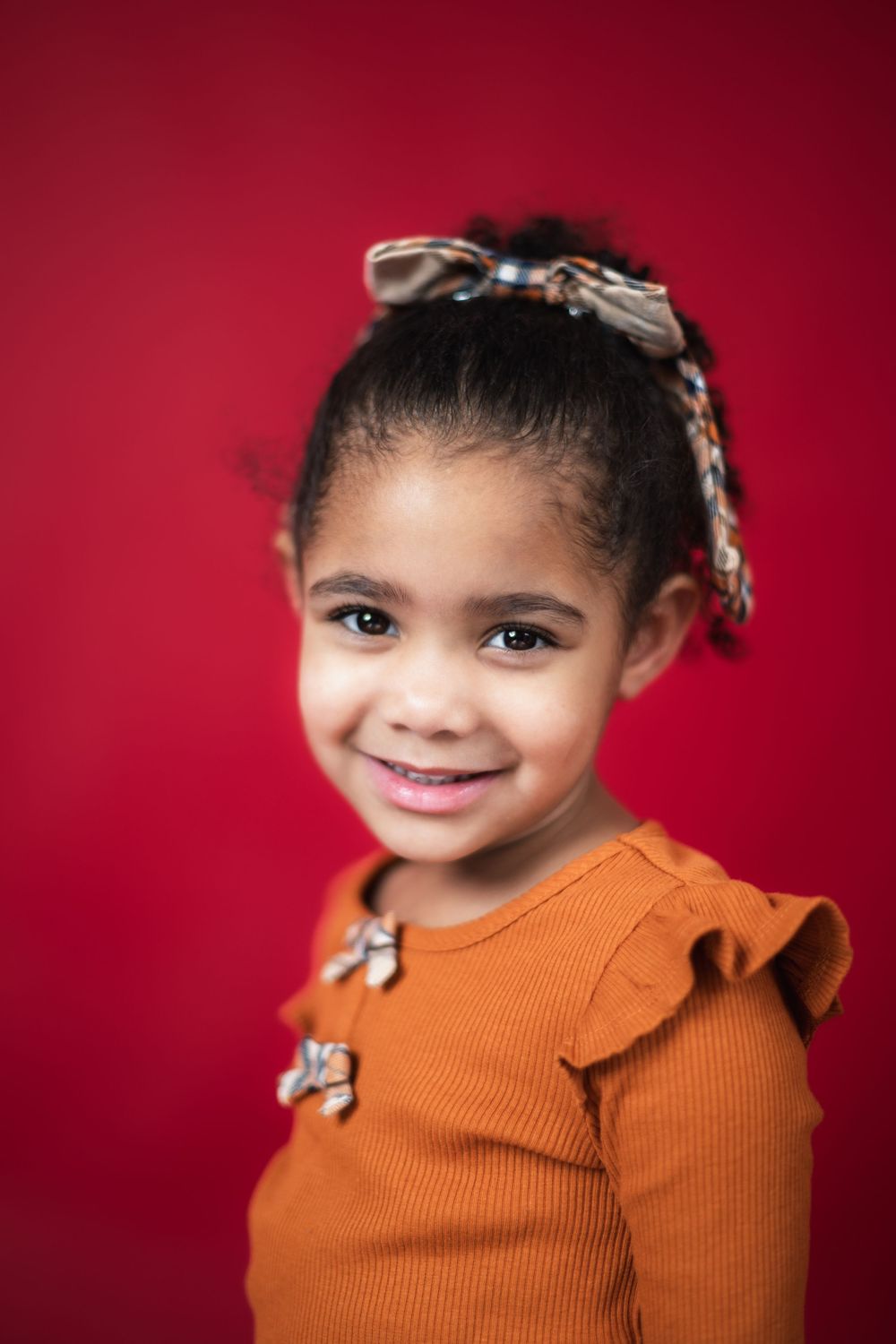 A child wearing an orange sweater smiles against a deep red backdrop in a series of connected portrait photos.