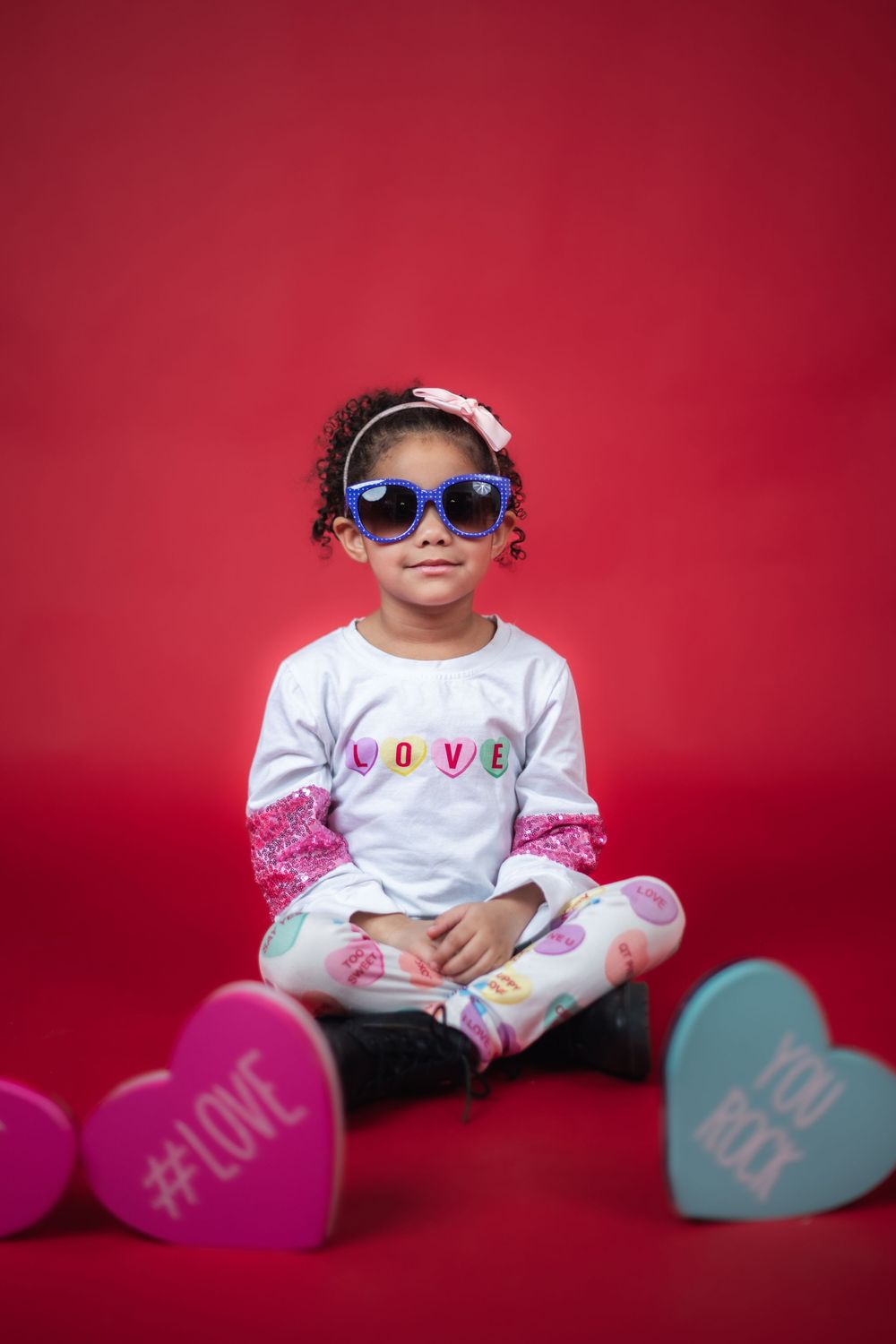 A small child wearing sunglasses and a white shirt poses with pink and blue heart decorations against a red backdrop.
