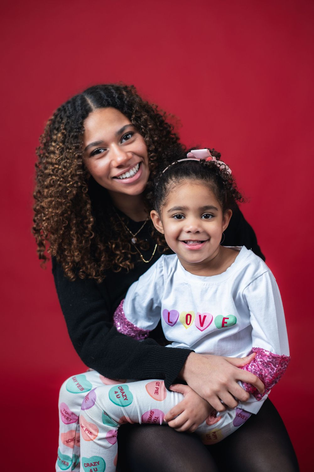 A joyful portrait session against a red backdrop shows warm embraces and genuine smiles in coordinated white outfits.