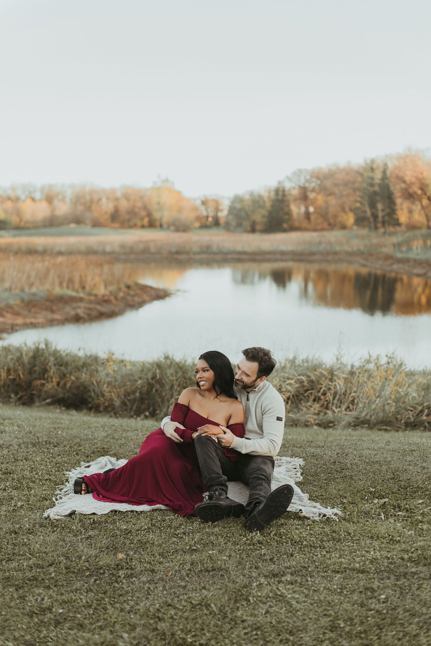 Couple sitting together on blanket by autumn pond, woman in flowing burgundy dress, romantic portrait.