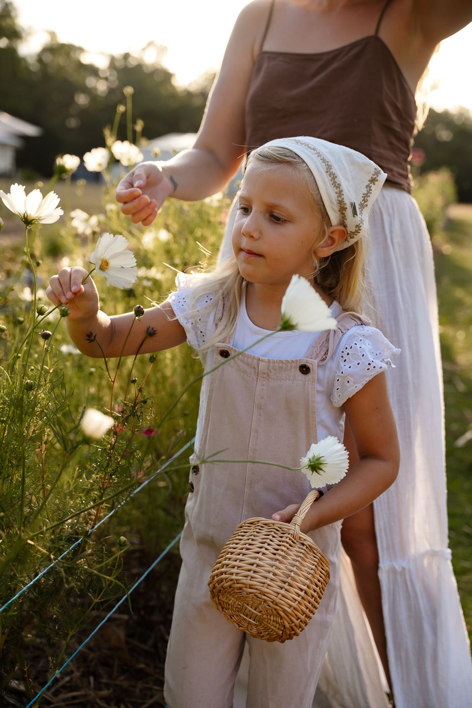 A child in beige overalls explores a wildflower meadow at sunset while holding a woven basket.