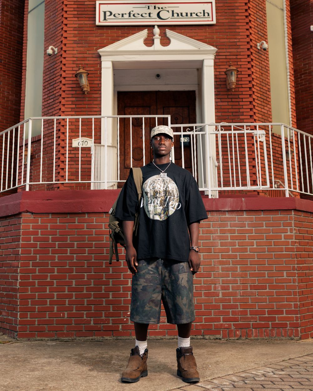 A person wearing baggy jeans and a dark shirt stands in front of a brick church building with white railings.