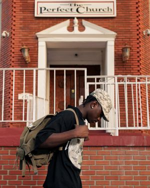 A person bows their head while standing in front of a red brick church building with a sign reading 'The Perfect Church'.