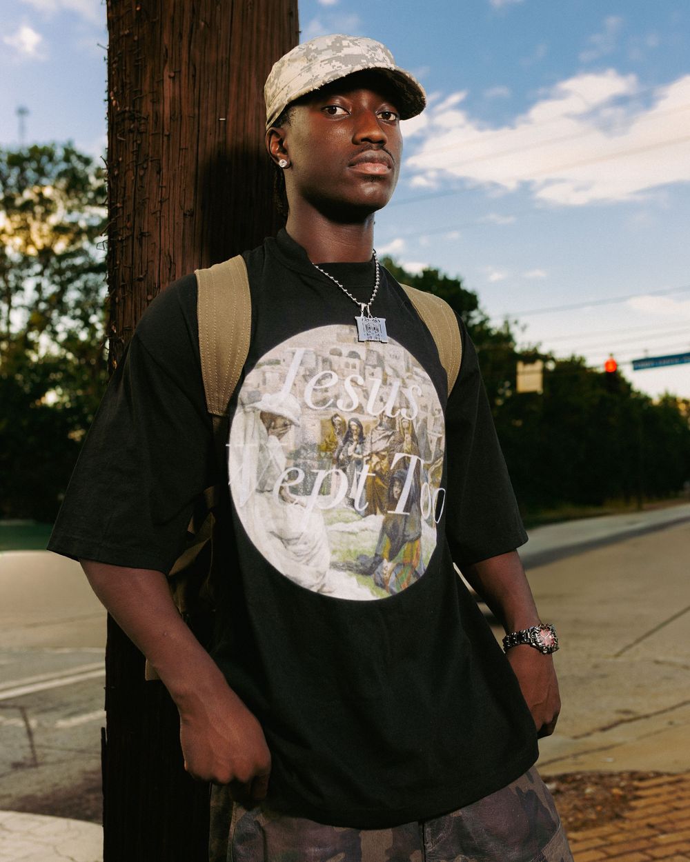 Person wearing a black t-shirt with a circular graphic design and a baseball cap stands near a wooden utility pole outdoors.
