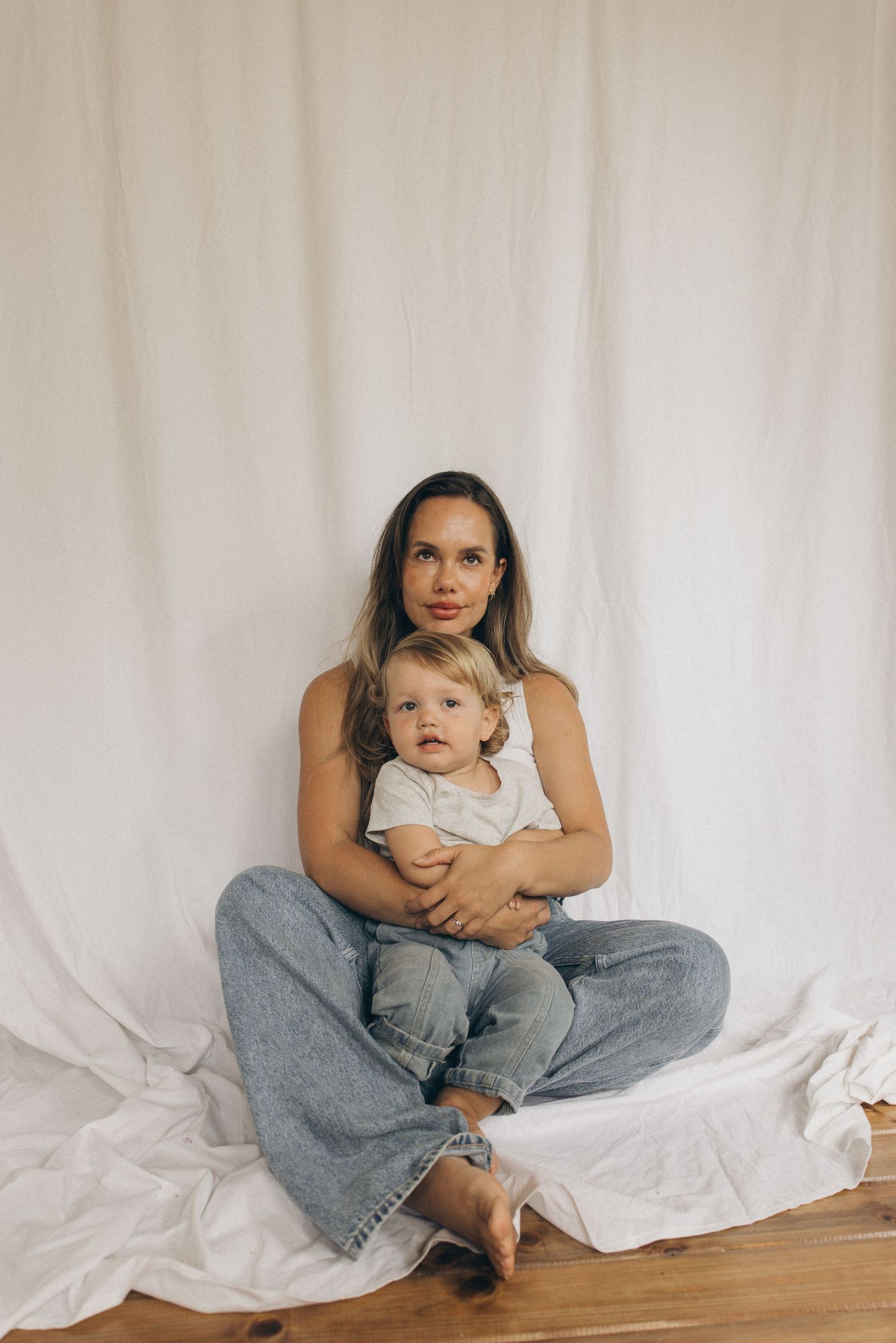 A mother in jeans and white top sits cross-legged holding her baby against a white backdrop in New Zealand.