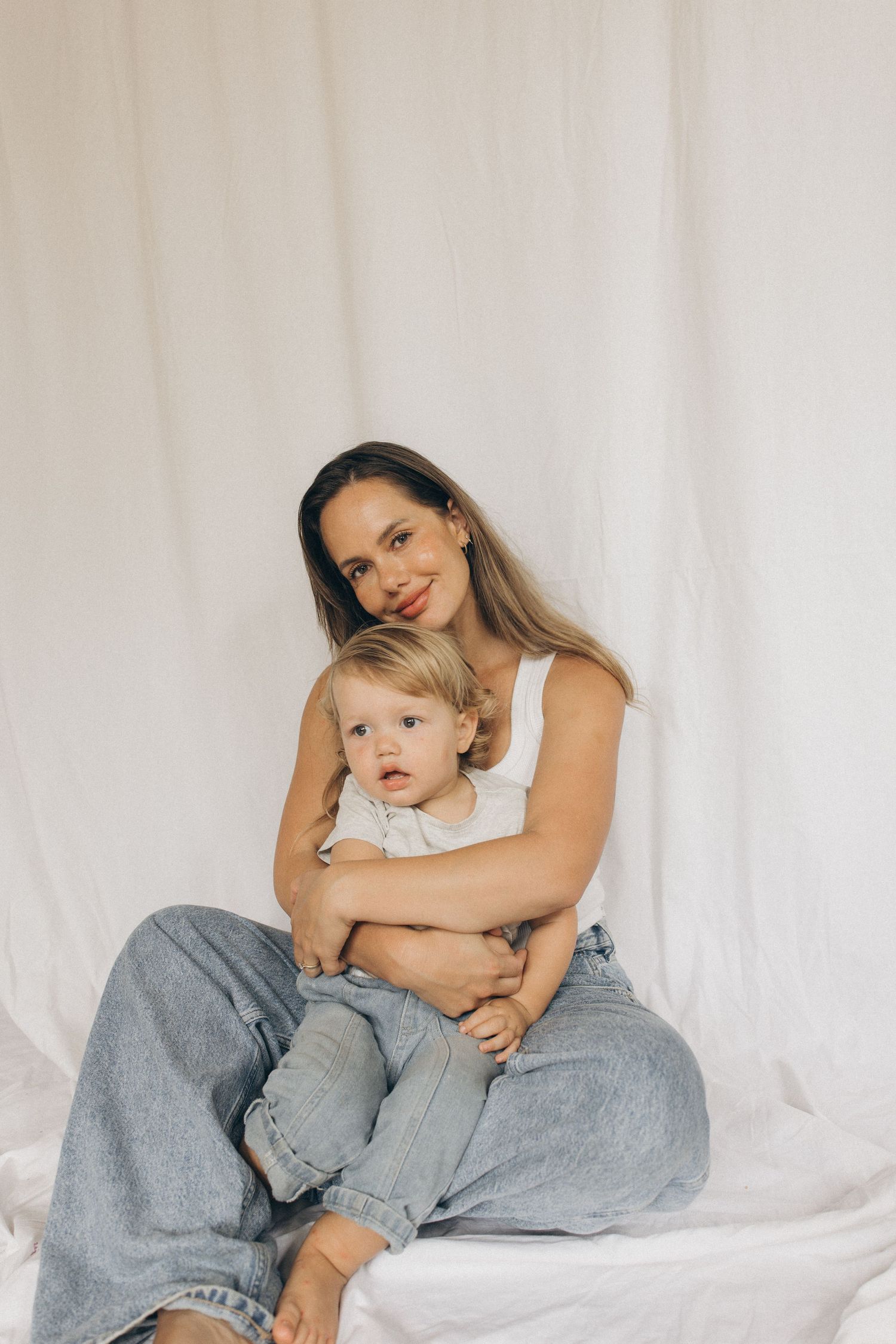A smiling mother in a white tank and jeans holds her toddler against a white fabric backdrop in New Zealand.