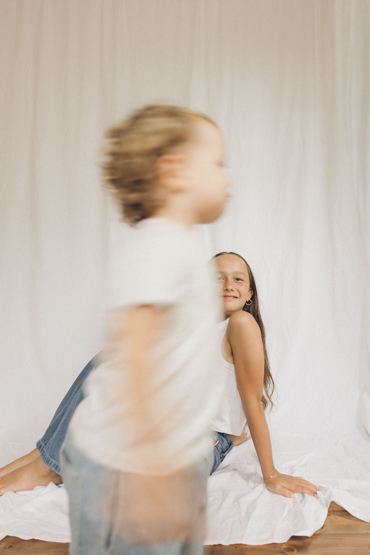 A smiling girl sits on a white backdrop while another child runs past in a blur in New Zealand.