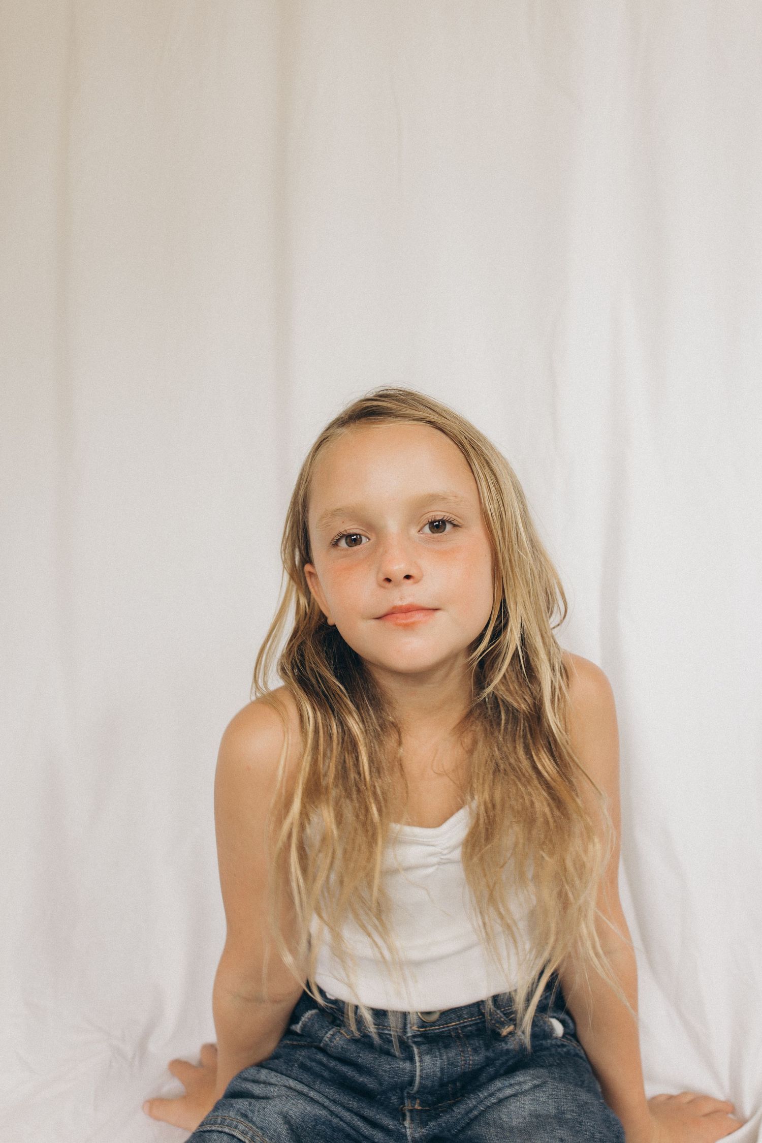 A young blonde girl in a white tank top and jeans poses against a white curtain backdrop in New Zealand.