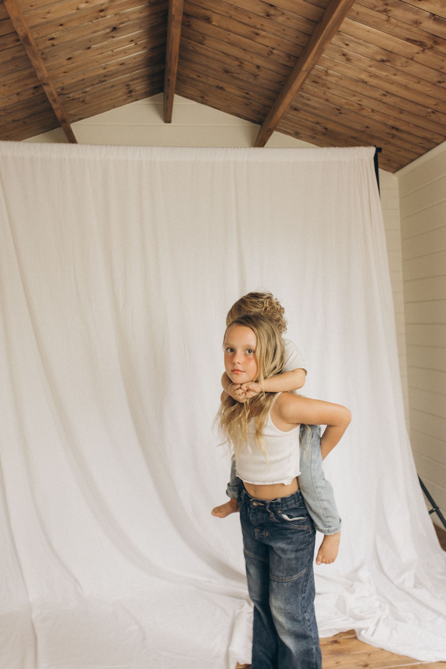 Young blonde girl in white crop top and jeans poses confidently in front of white fabric backdrop in wood-beamed studio in New Zealand.