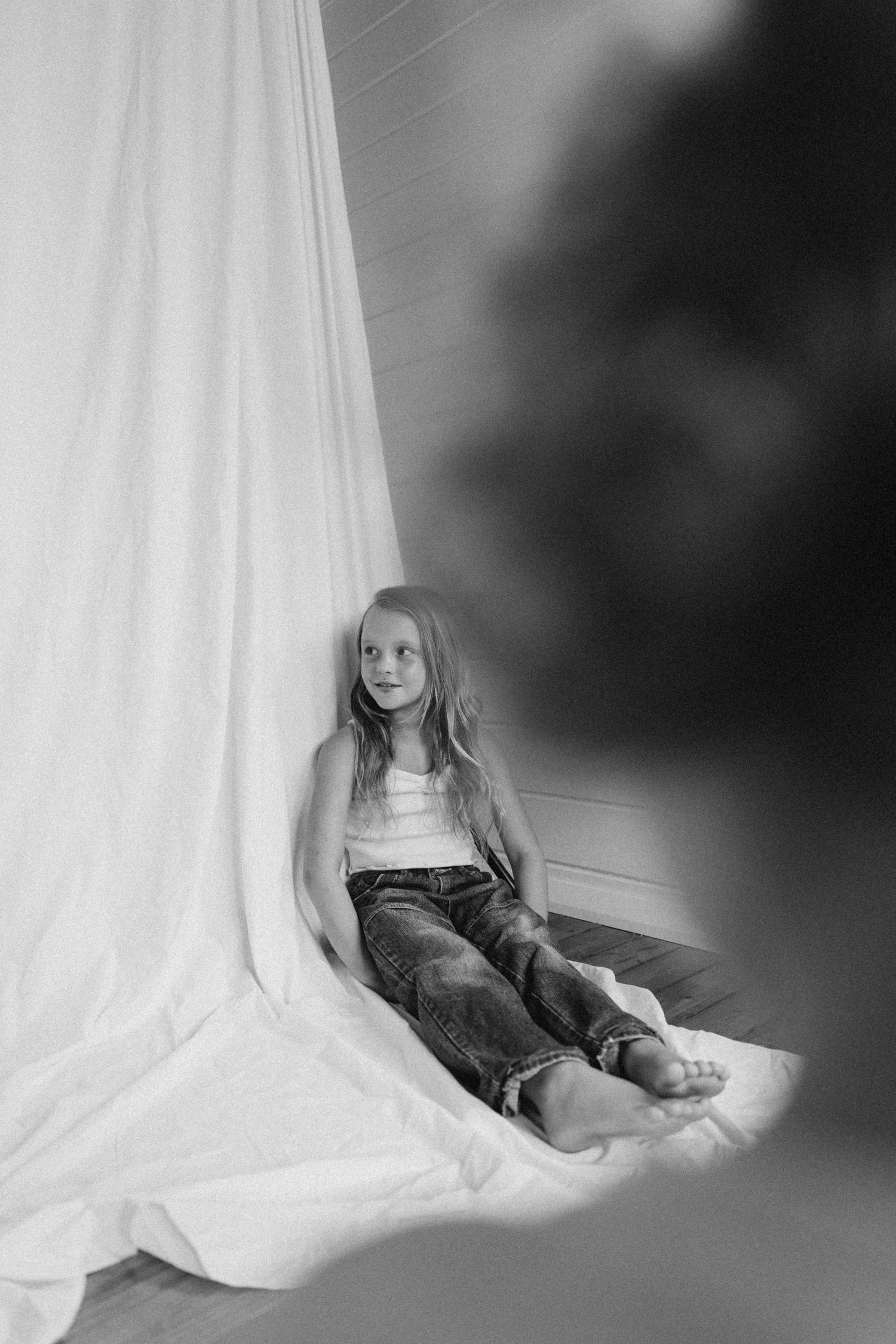 Black and white photo of a young girl in a white tank top and jeans sitting against a white curtain backdrop in New Zealand.