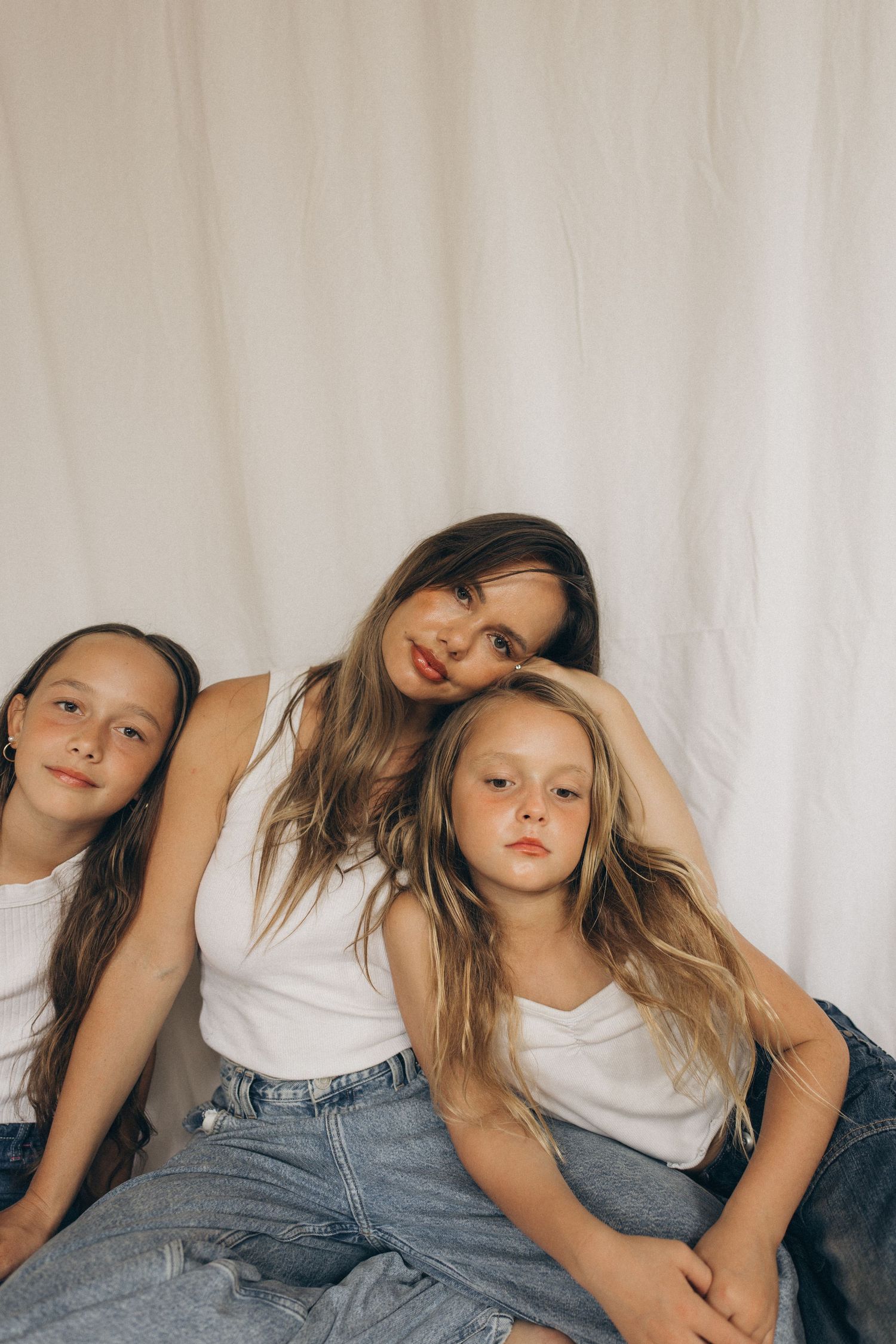 A mother leans between her two daughters, all wearing white tops and jeans against a white fabric backdrop in New Zealand.