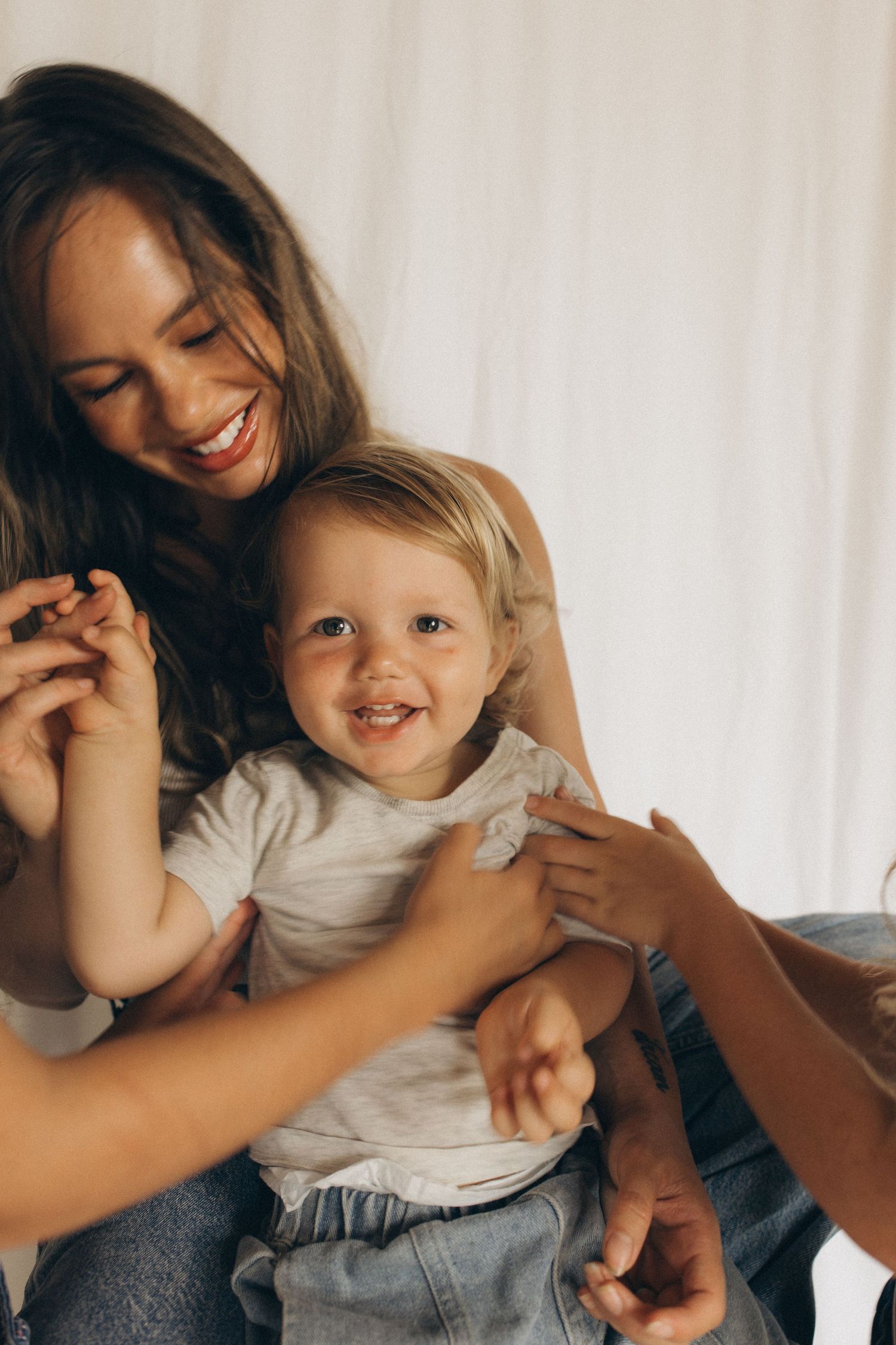 Smiling mother holds laughing toddler in white shirt while another person reaches in to play with the happy child in New Zealand.