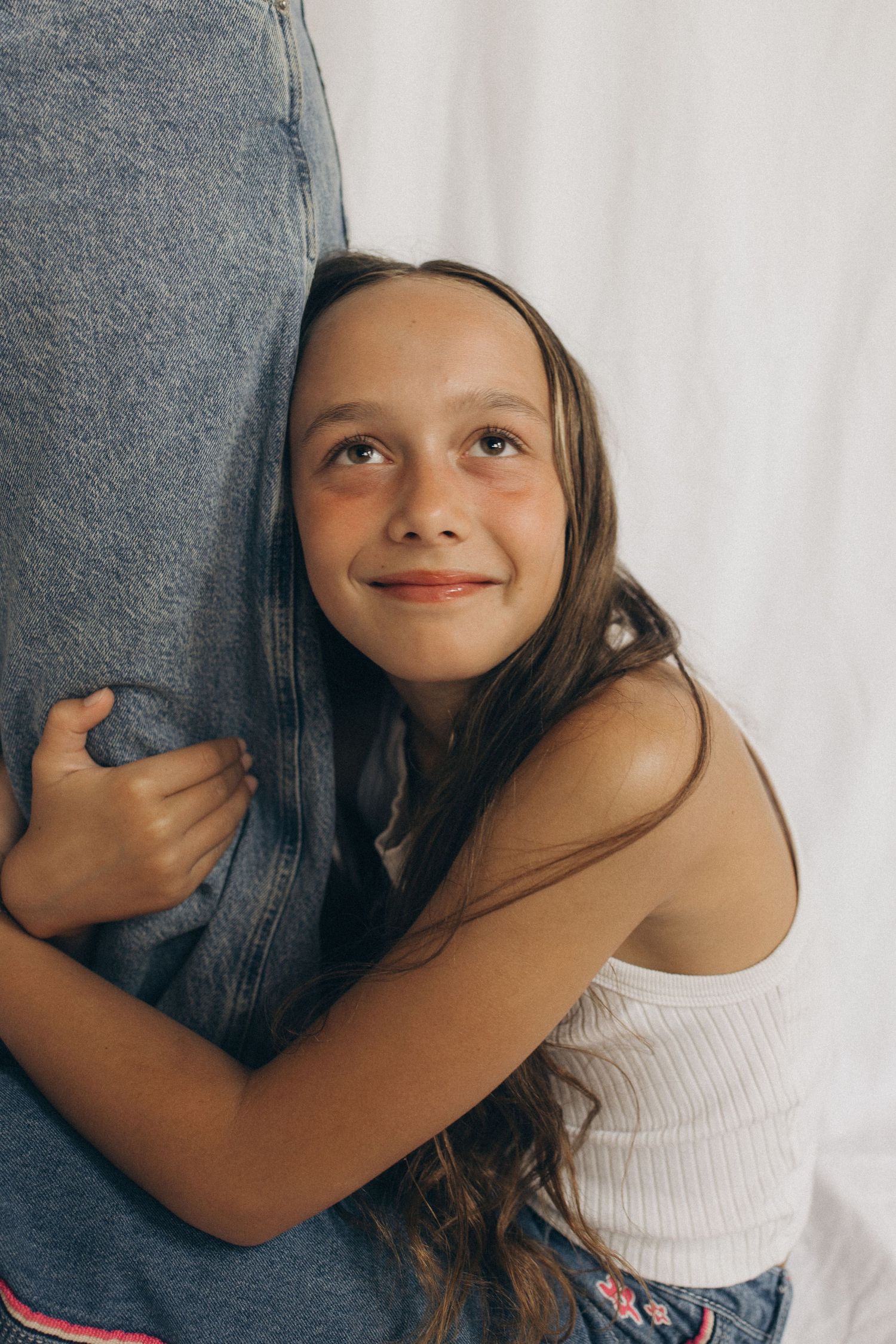 A smiling young girl with long brown hair hugs an adult while looking up, wearing a white tank top in New Zealand.