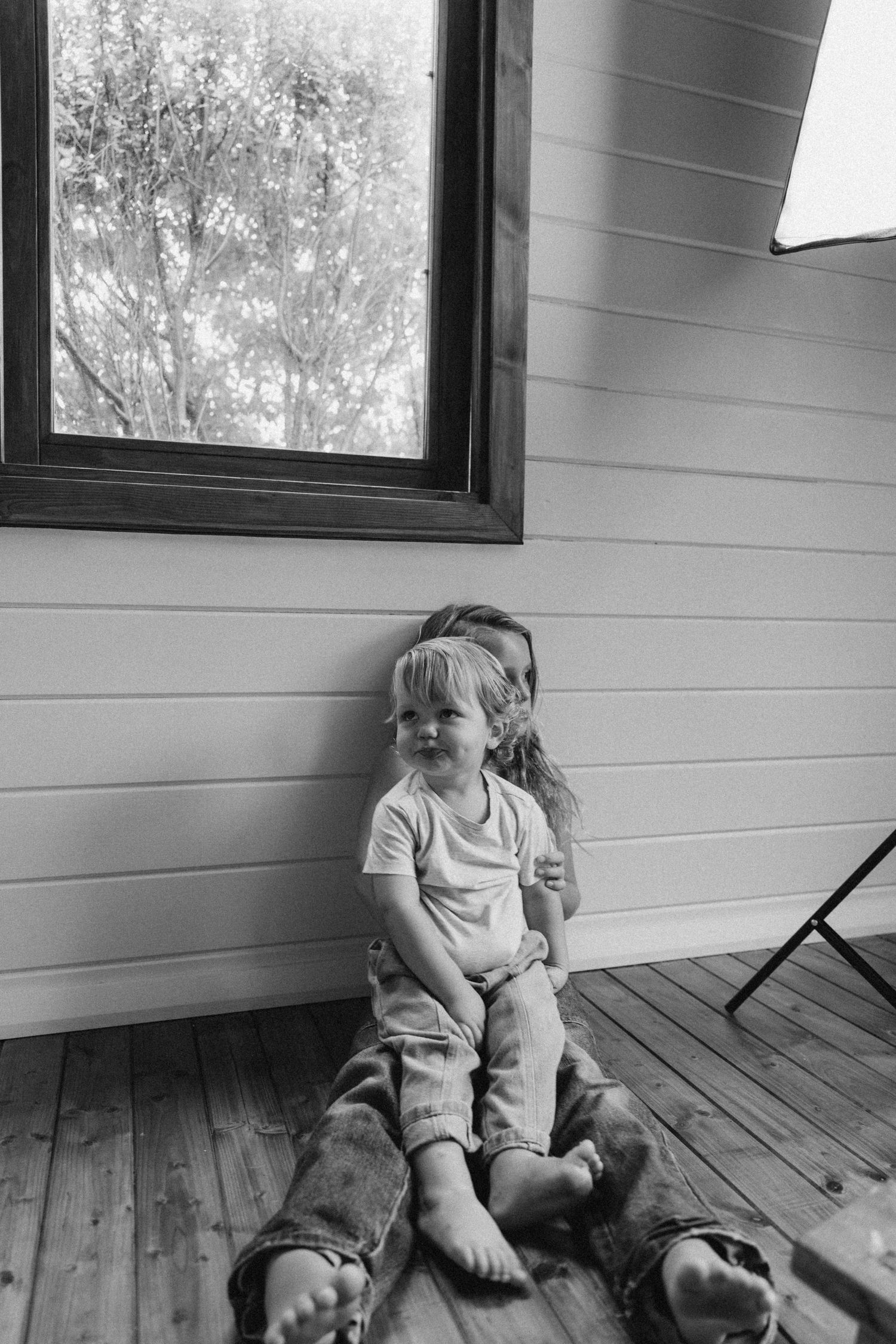 Black and white photo of a toddler sitting on a wooden floor with an older child behind them near a window in New Zealand.