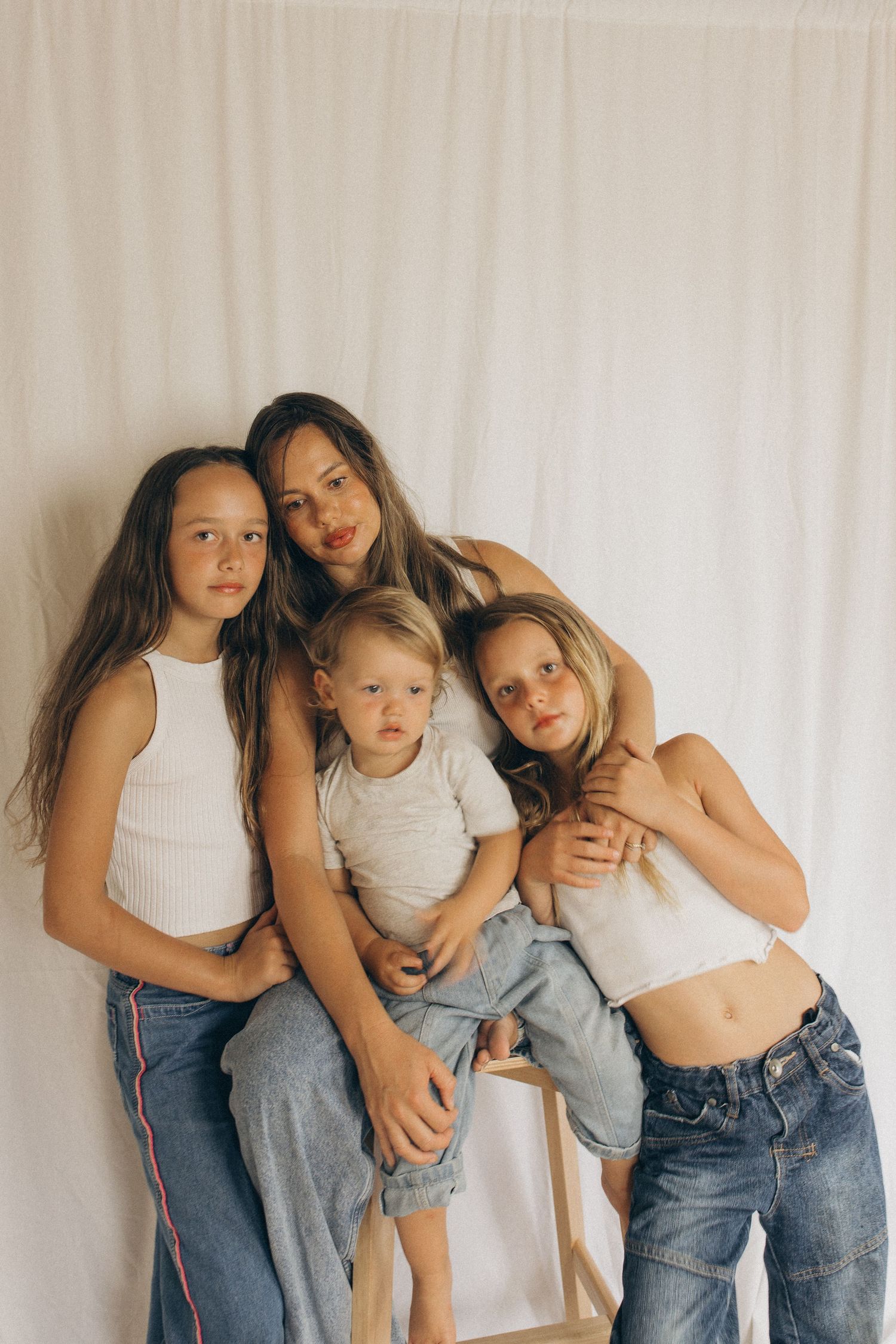 A mother and her three children pose together in white tops and jeans against a white curtain backdrop in New Zealand.