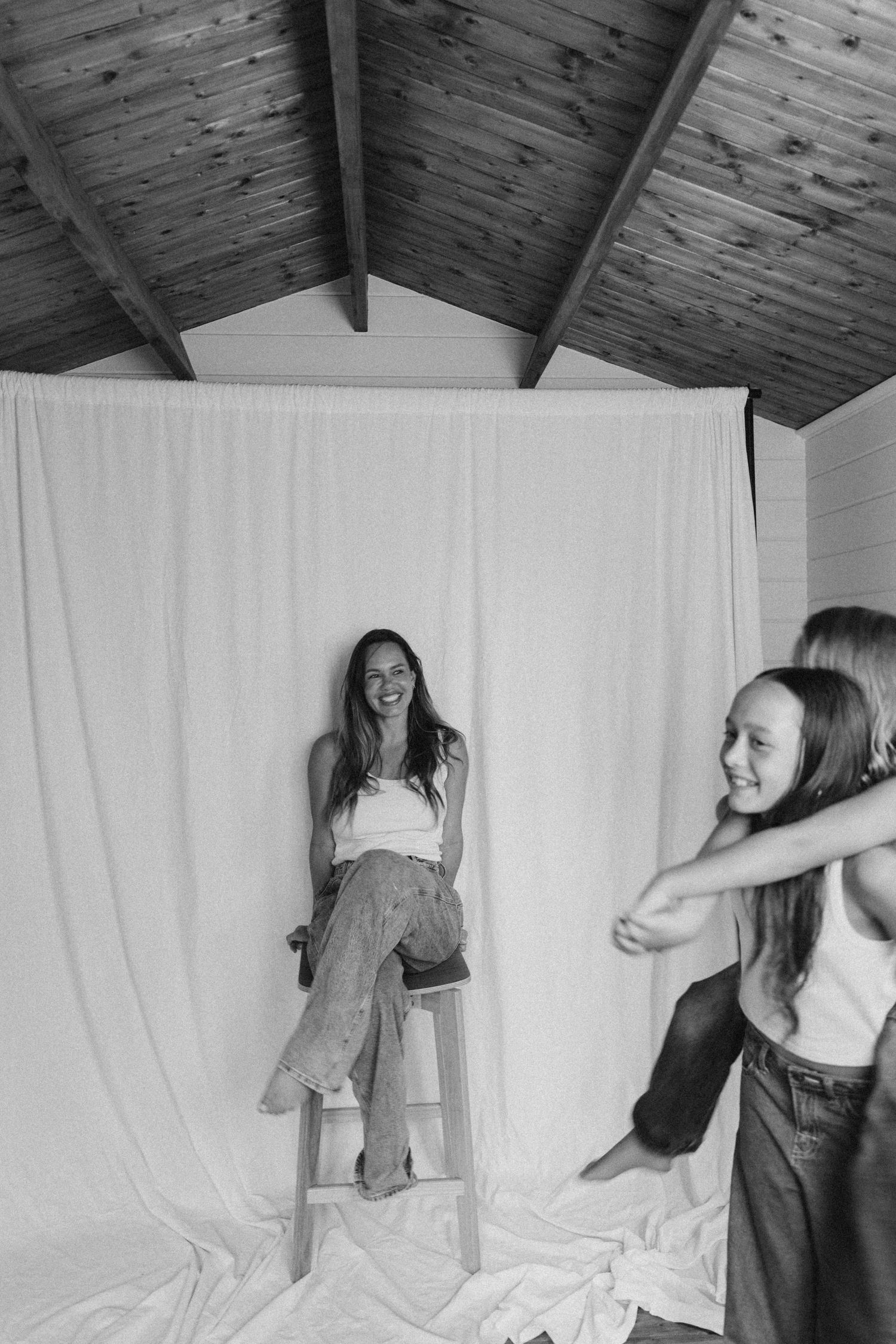 Black and white photo of a woman sitting on a stool laughing while two girls play in front of a white backdrop in New Zealand.