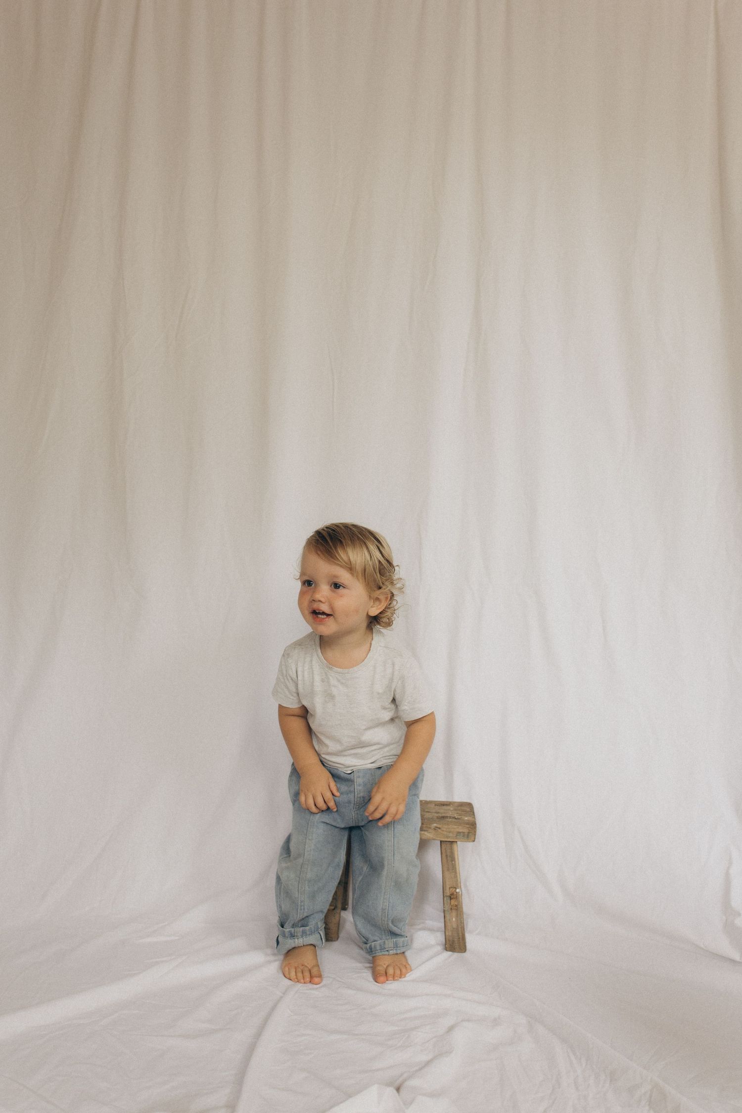 A smiling toddler in a grey t-shirt and jeans stands barefoot next to a wooden stool against a white backdrop in New Zealand.