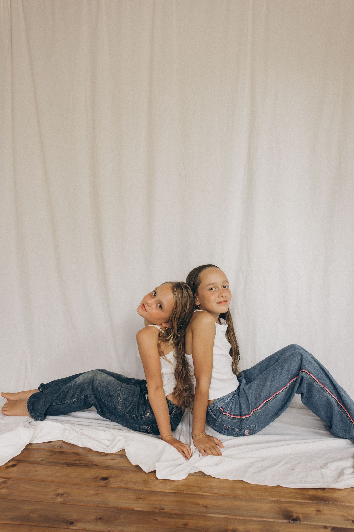 Two girls in white tanks and jeans sit back-to-back on a white backdrop on a wooden floor in New Zealand.