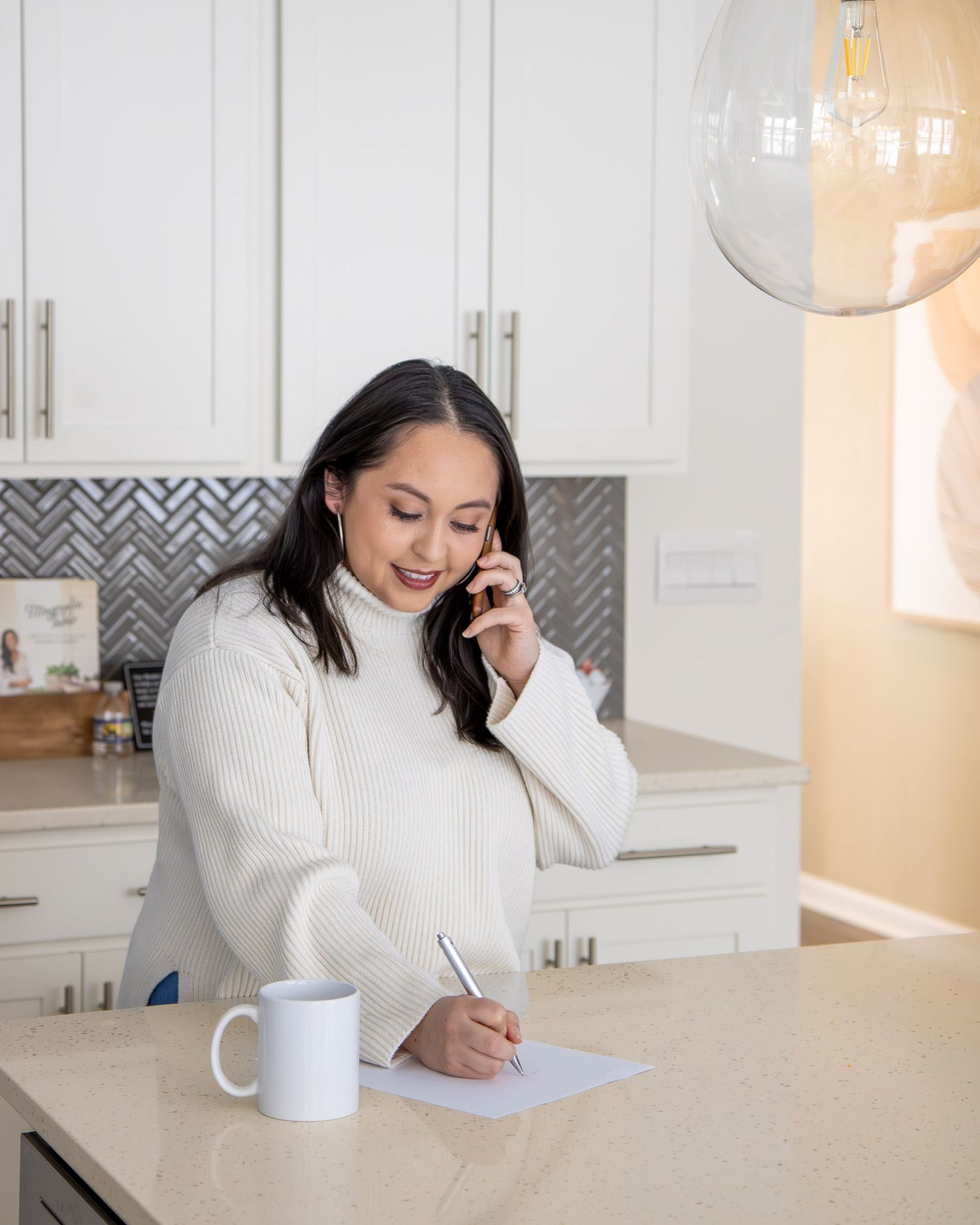 A realtor in a white sweater works at modern kitchen counter with laptop and paperwork in bright home office setting.