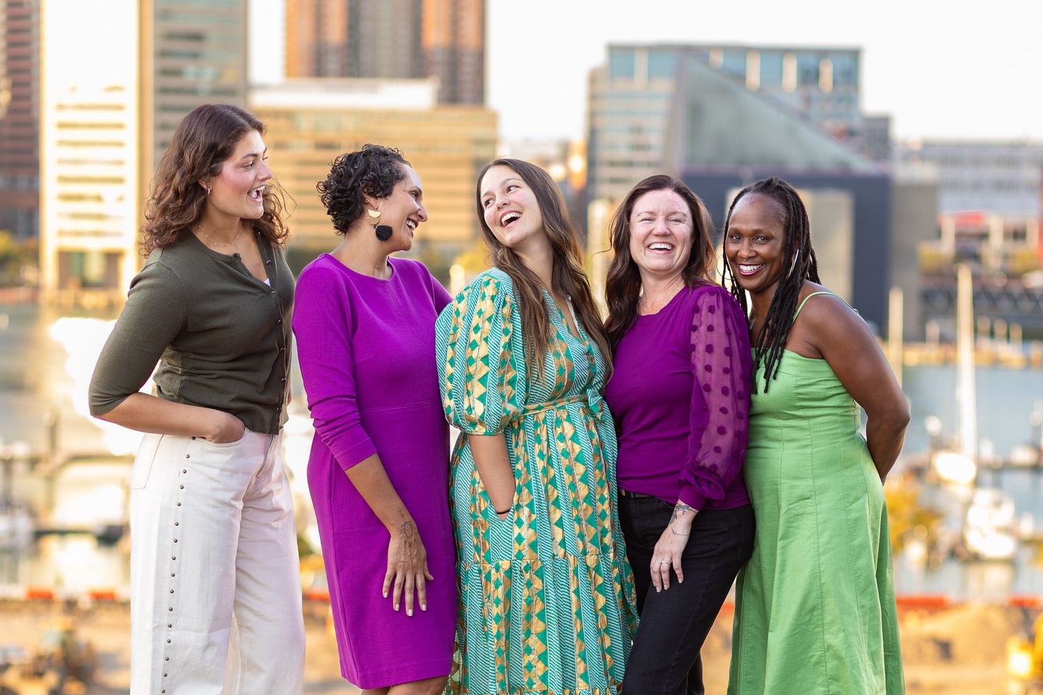 A group of professional women wearing coordinated green and purple dresses laugh together at the Inner Harbor in Baltimore.