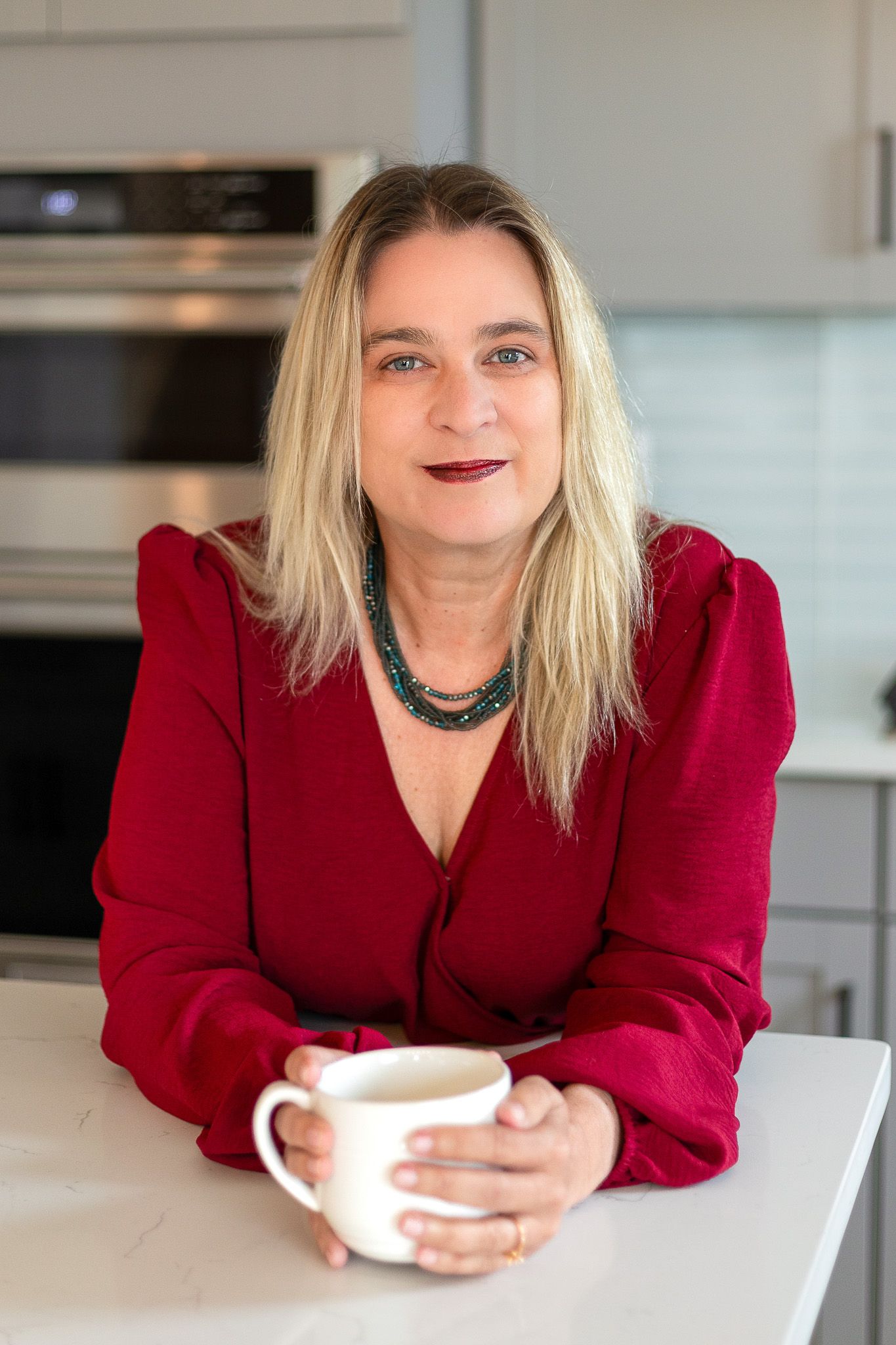 Woman in red pantsuit leaning across a white table holding coffee cup in modern kitchen setting.