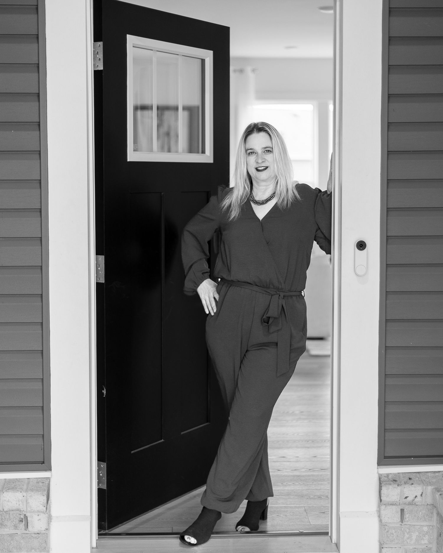 A black and white personal branding photo of a realtor posing in a doorway wearing a v-neck jumpsuit with a belted waist.