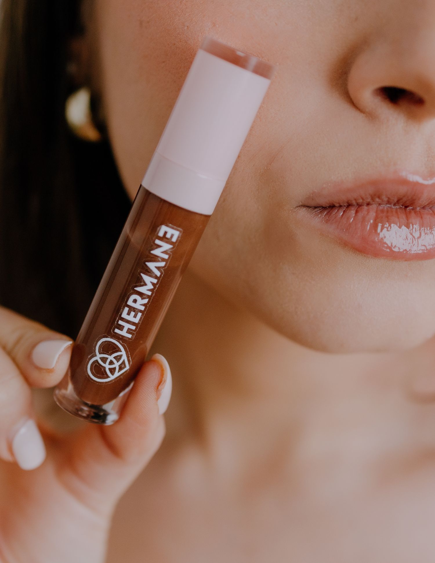 Close up of a hand holding a Her Mane lip gloss product with glossy lips visible in background.