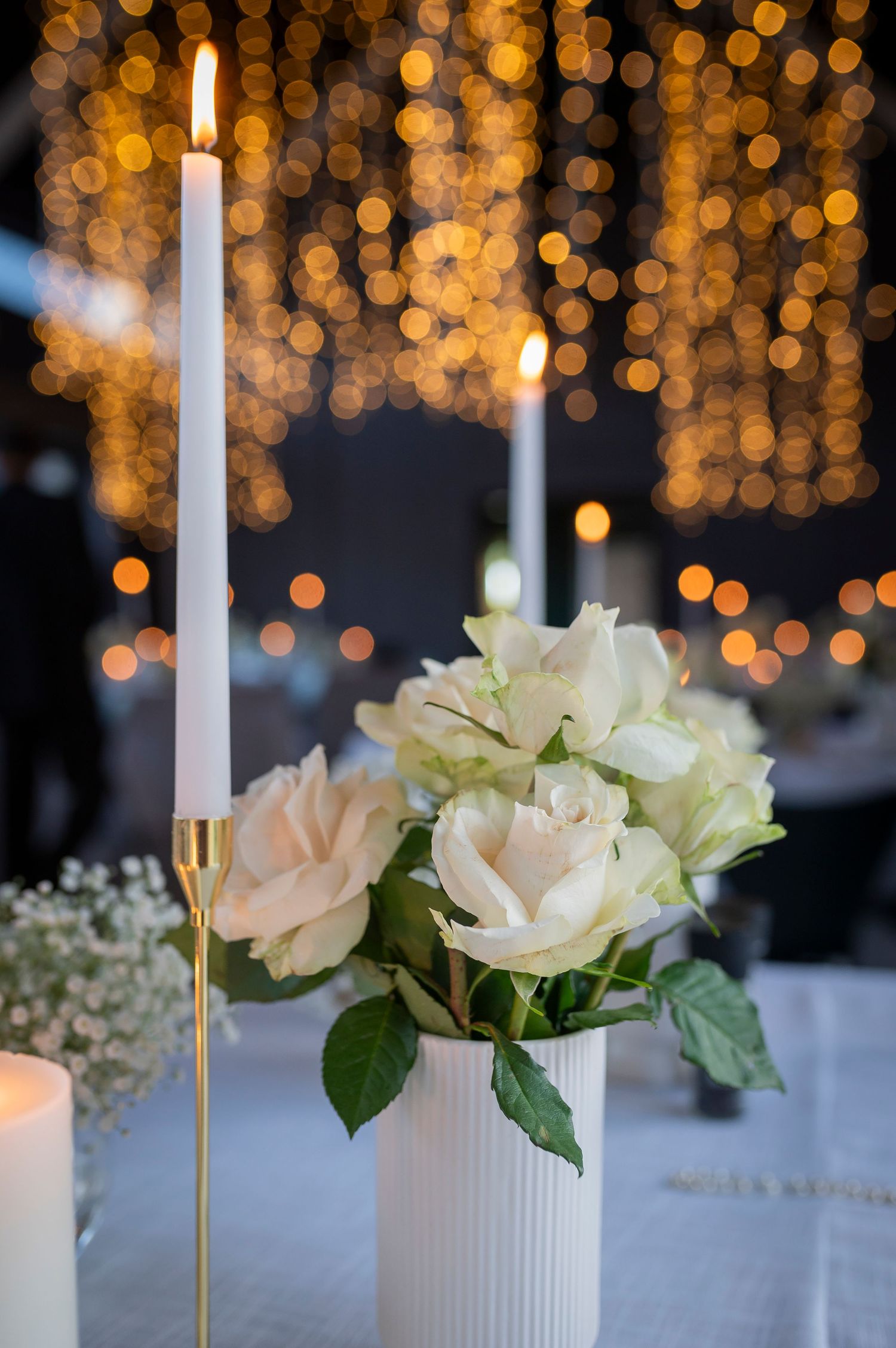 White roses in ceramic vases with tall white candles against a backdrop of glowing golden bokeh lights create an elegant tablescape.