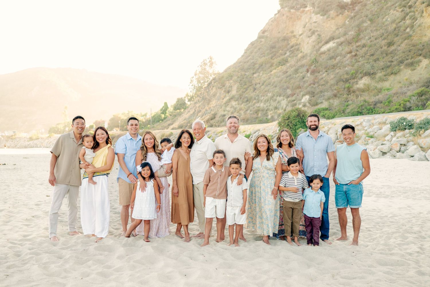 A group of people in coordinated beige and white outfits standing together on a sunny beach with cliffs in the background.