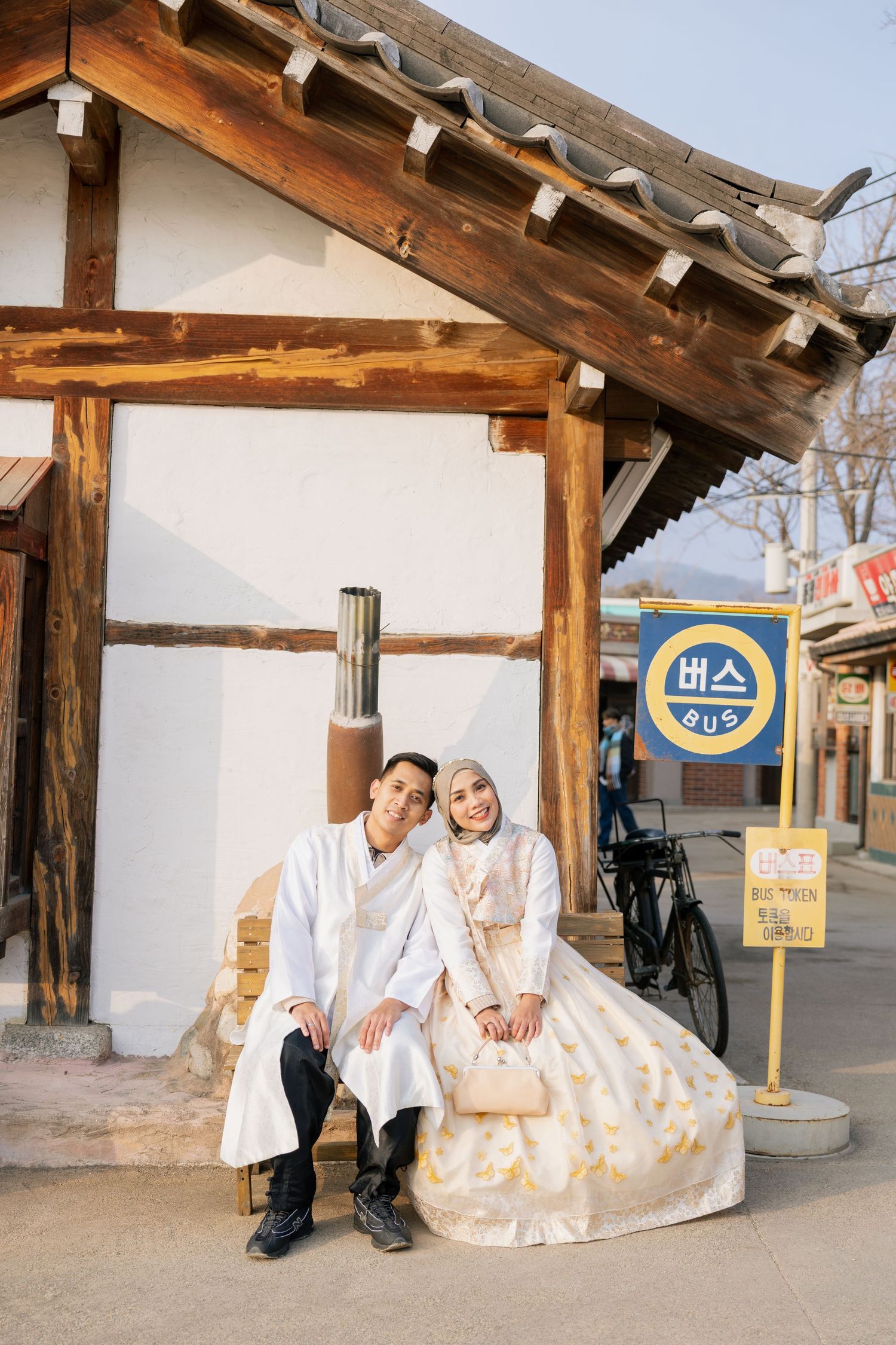 Traditional Korean hanbok wedding attire photographed at a rustic outdoor location with wooden architecture and white walls.