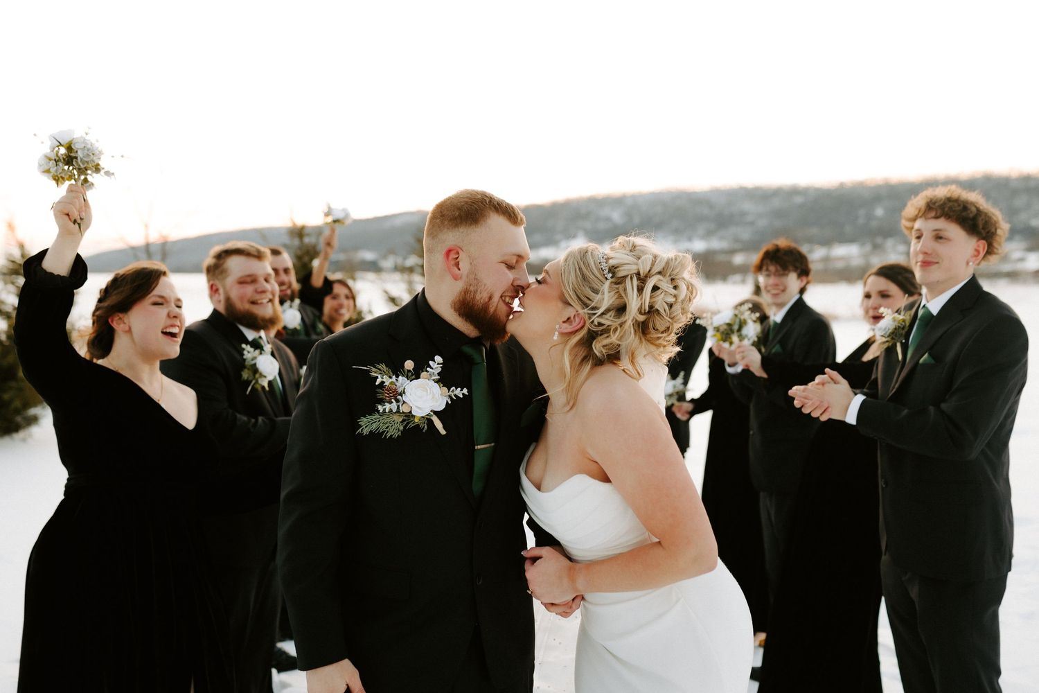 Wedding party celebrates with joyful couple kissing in snowy winter landscape.