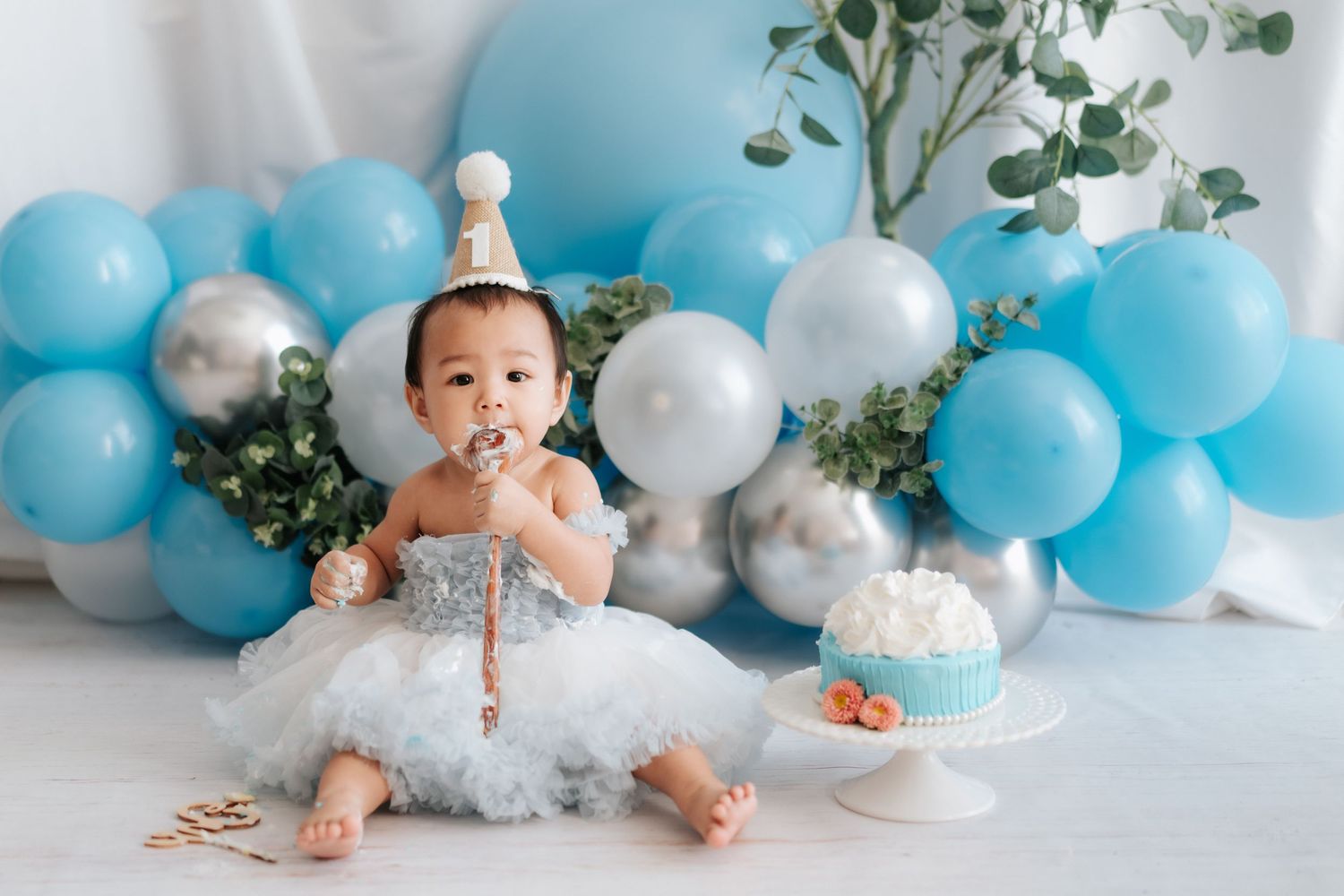 A baby sits in a white chair surrounded by floral decorations and balloons at a birthday celebration.