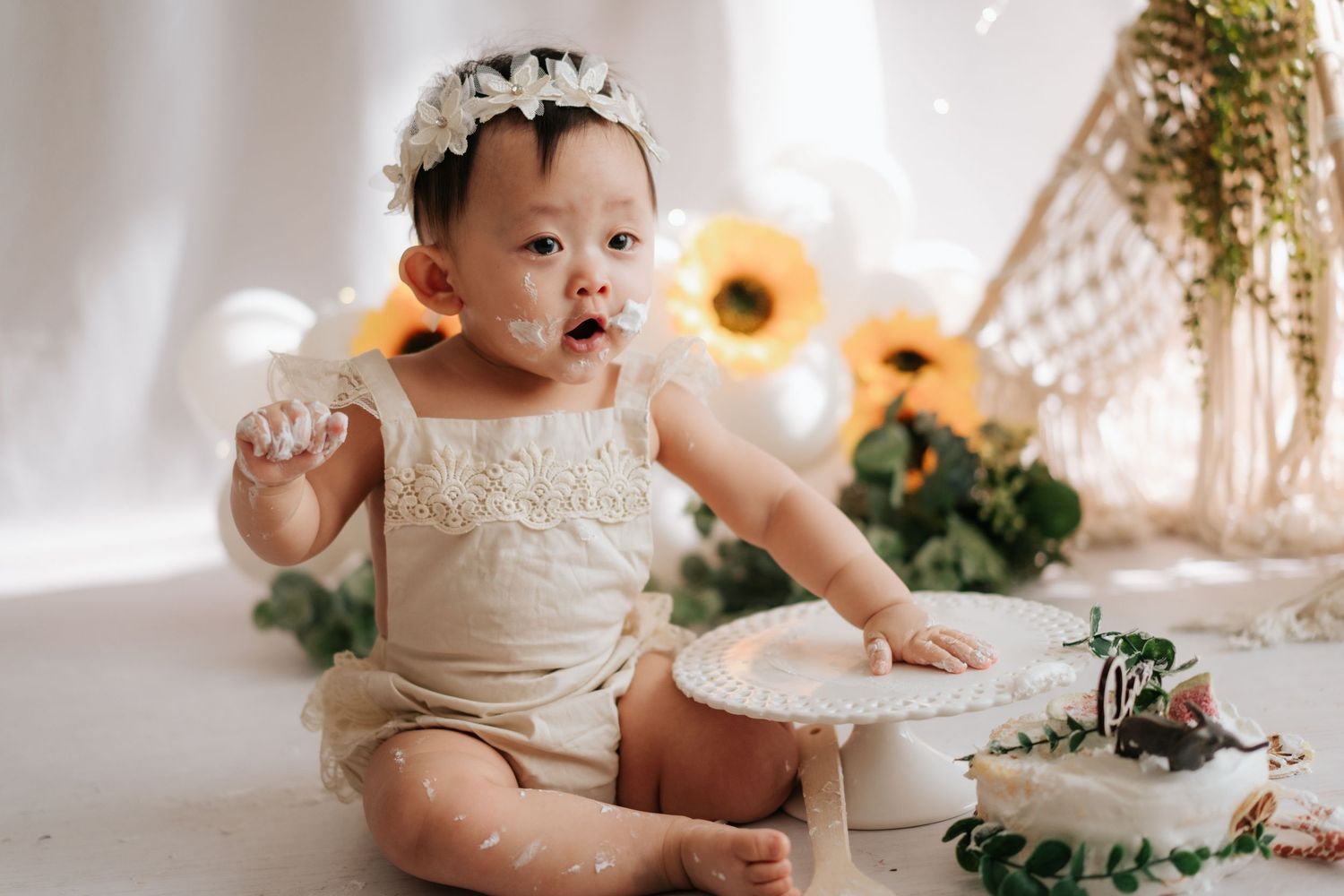 A baby in a party hat sits on a wood slice with white balloons and greenery decorations in the background.