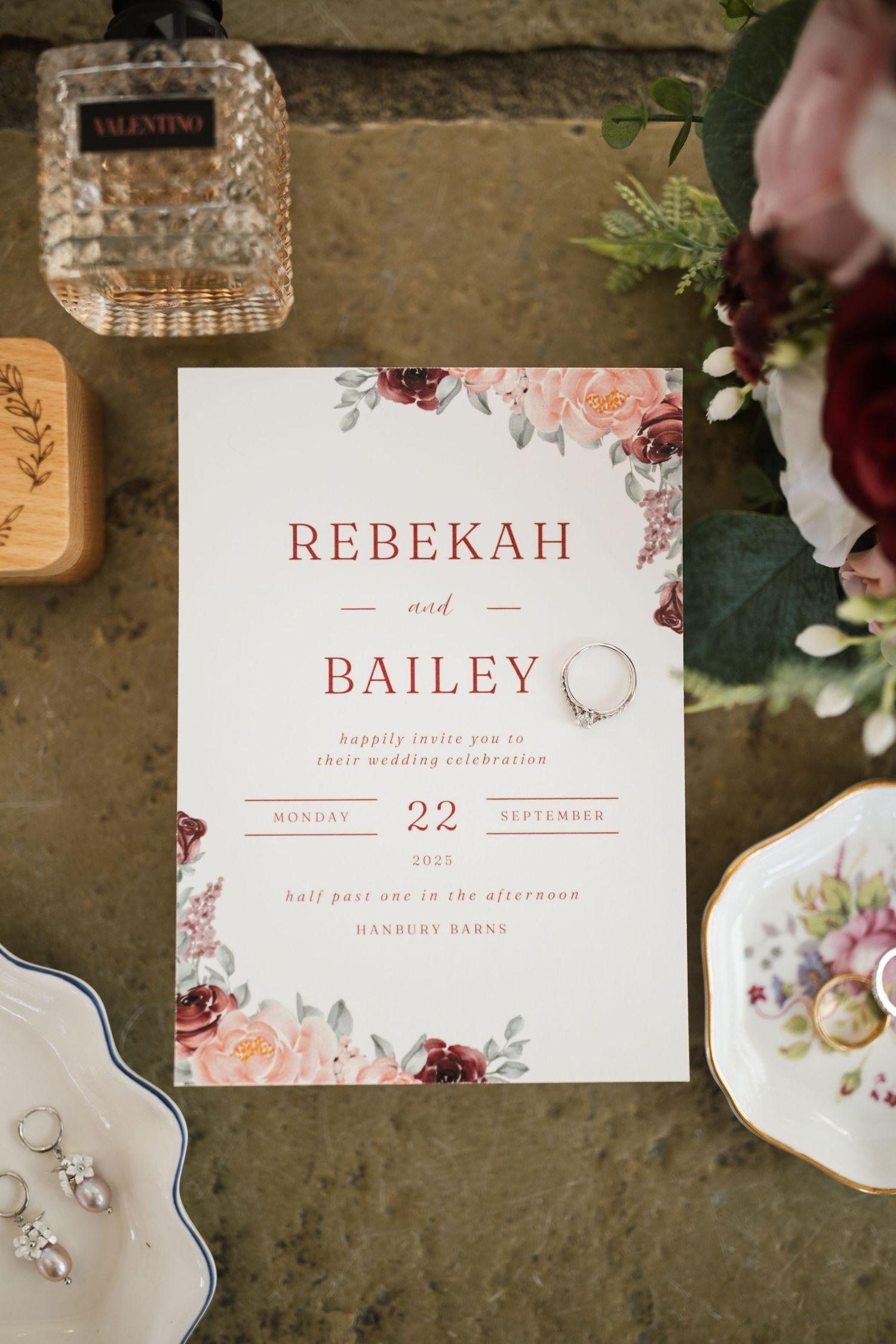 Wedding invitation suite with floral designs and burgundy accents displayed with perfume bottle and jewelry on textured surface.