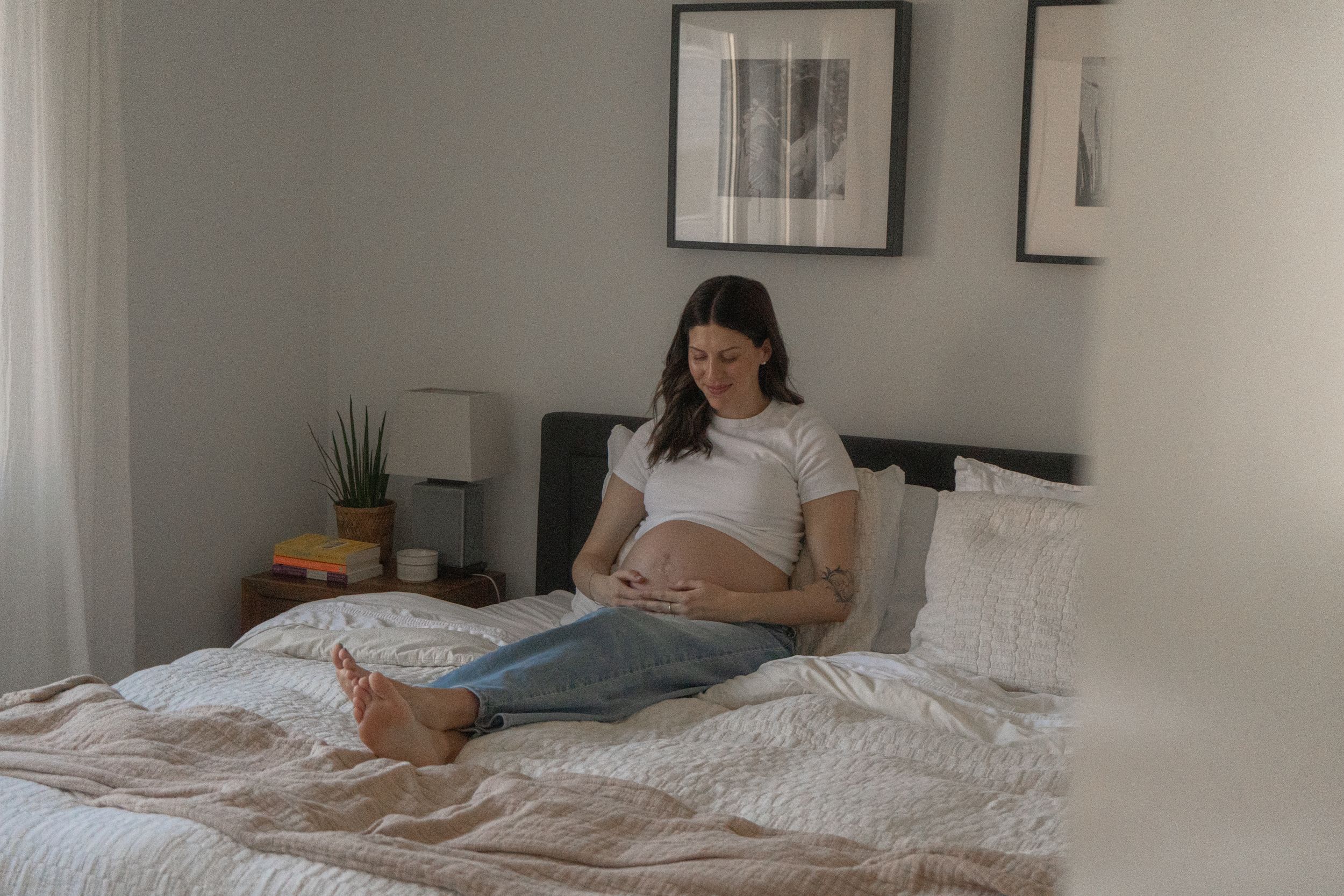 Pregnant woman sitting on bed cradling baby bump, wearing white t-shirt and jeans in cozy bedroom.