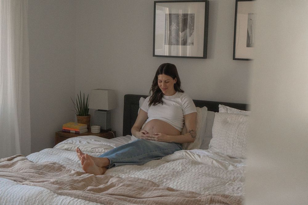 Pregnant woman sitting on bed cradling baby bump, wearing white t-shirt and jeans in cozy bedroom.