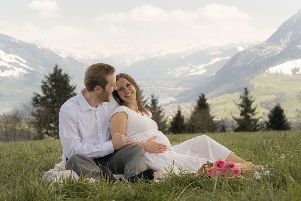 Pregnant couple sitting on a grassy alpine meadow with snowy mountains in the background on a cloudy day.